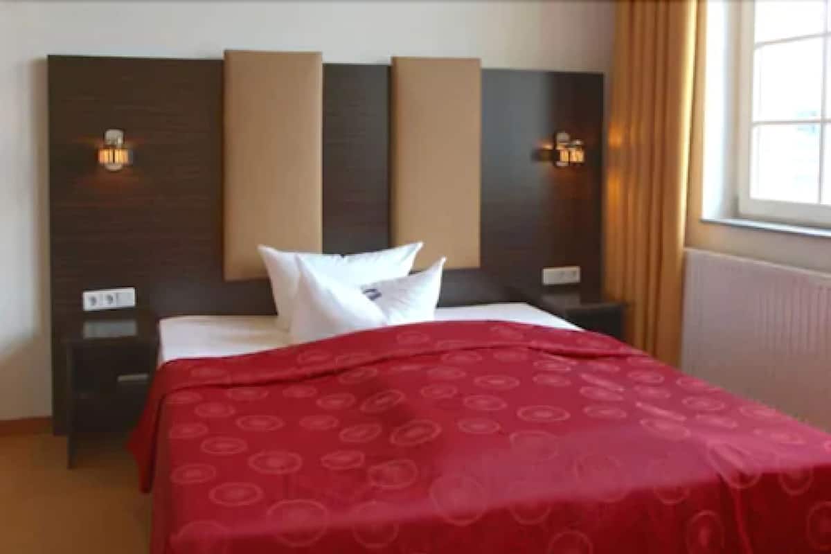 A hotel room with a bed, two bedside tables, a nightstand, a window with curtains, and wall-mounted lighting.