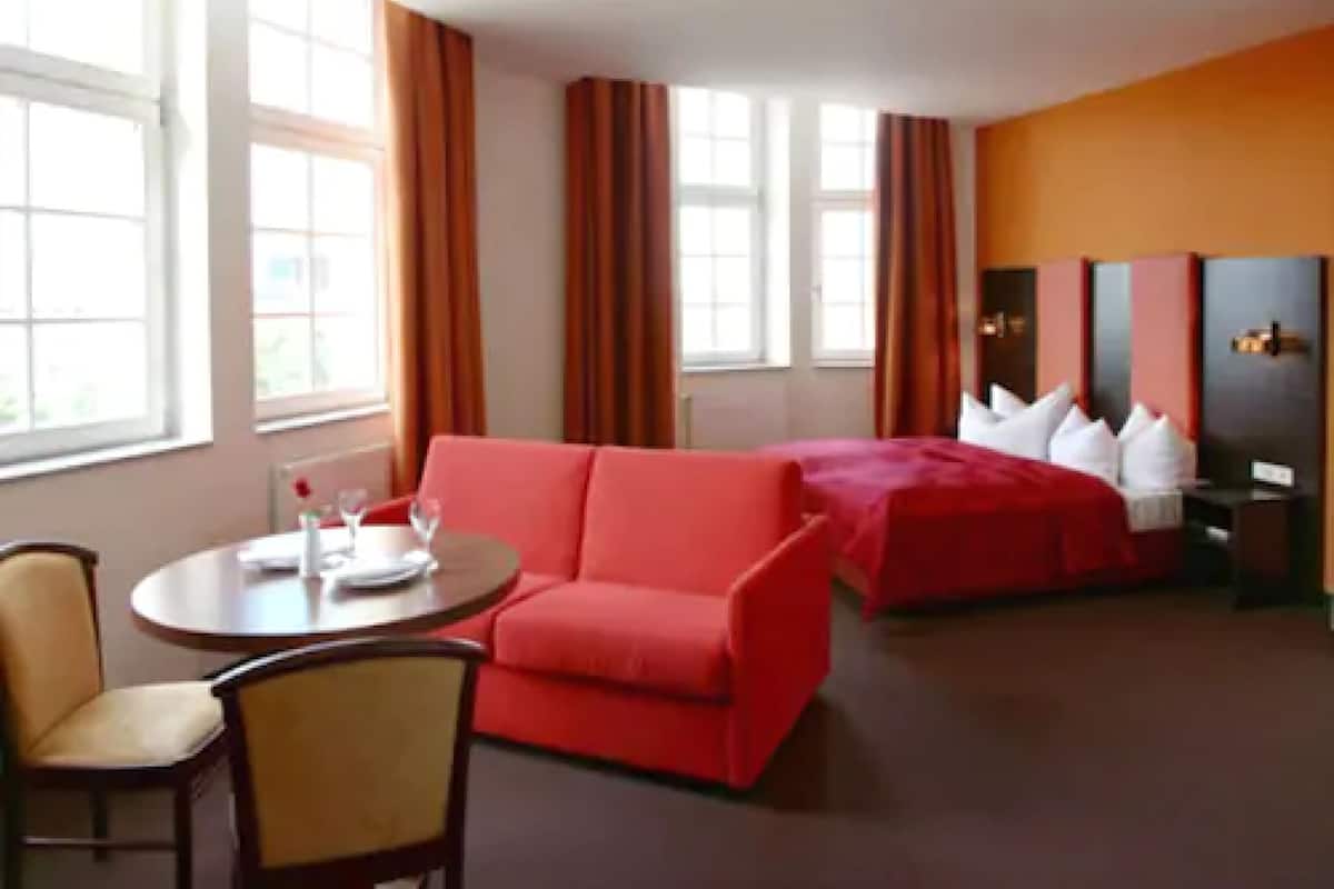 A hotel room with a red sofa, a bed with white pillows, a small table with a glass of water, and a nightstand.