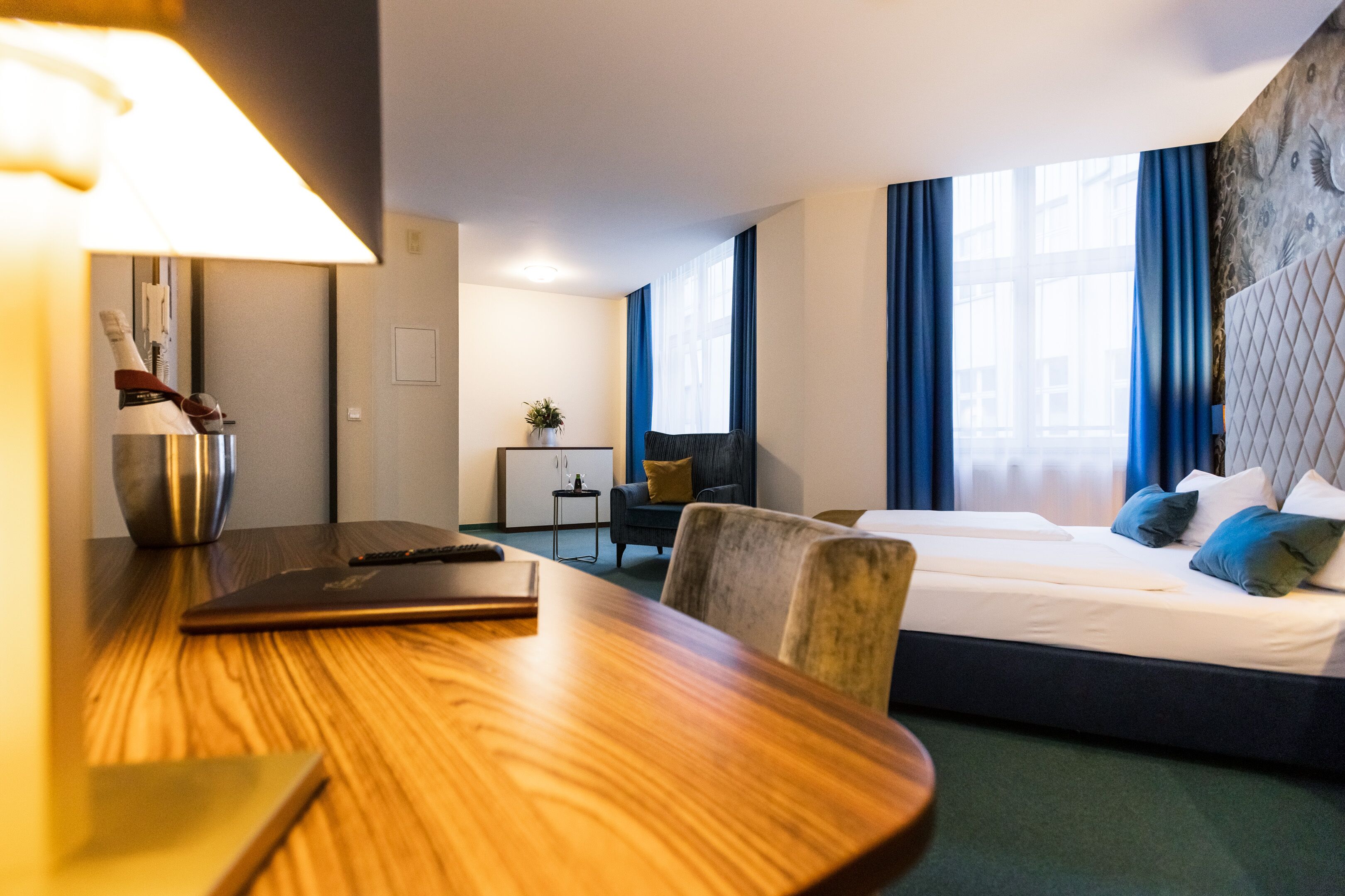 Ein Hotelzimmer mit einem Holztisch, einem Bett mit blau-weißer Bettwäsche, einem Nachttisch und einem großen Fenster mit blauen Vorhängen.