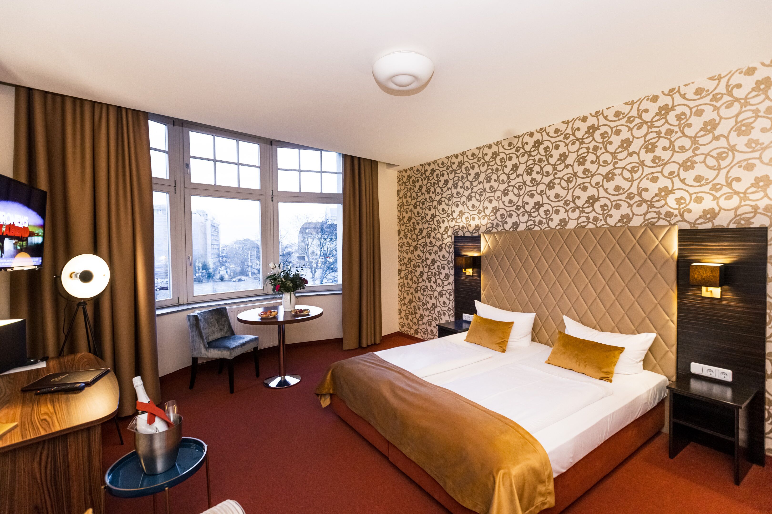 A hotel room with a large bed, a desk, a chair, a small table with a vase, and a window with a city view.