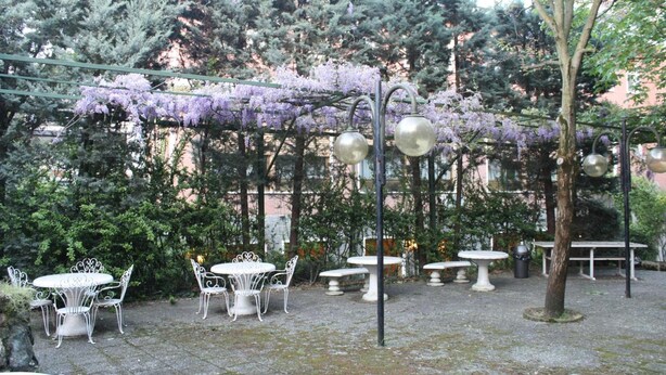 Zona exterior de terrassa amb taules i cadires rodones, envoltada de vegetació i enfiladeres de flors morades.
