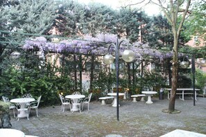 Ein Außenbereich mit einer Terrasse, ausgestattet mit runden Tischen und Stühlen, umgeben von Pflanzen und blühenden, violetten Weinreben.