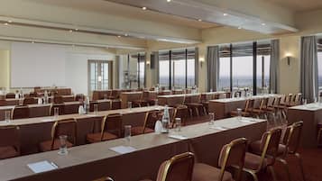 Ein Konferenzraum mit mehreren Tischen und Stühlen, großen Fenstern mit Meerblick und einer Projektionsleinwand.