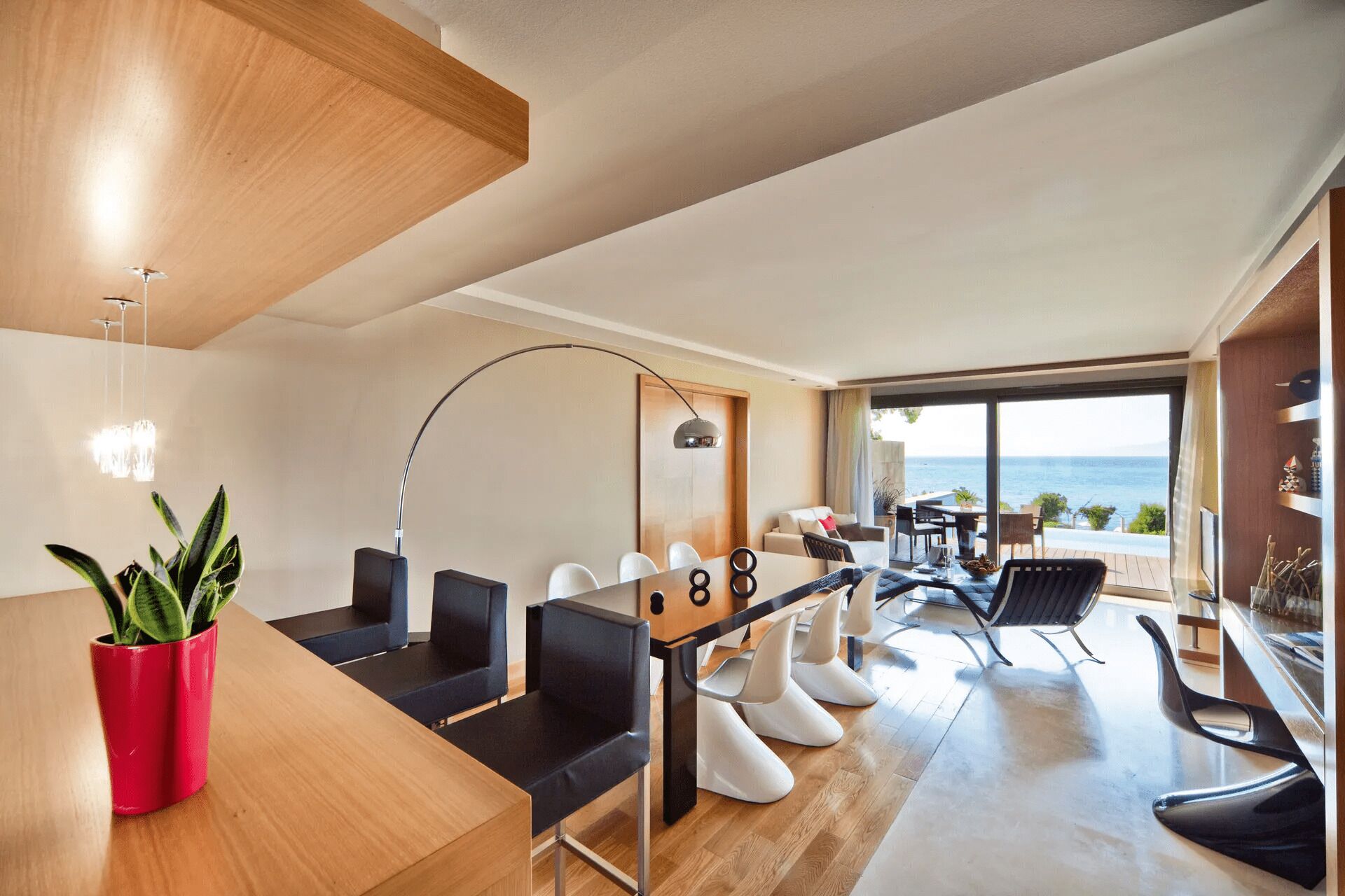 Una habitación de hotel moderna con techo de madera, un área de comedor con mesa y sillas, un ventanal con vistas al mar y un balcón con mobiliario exterior.