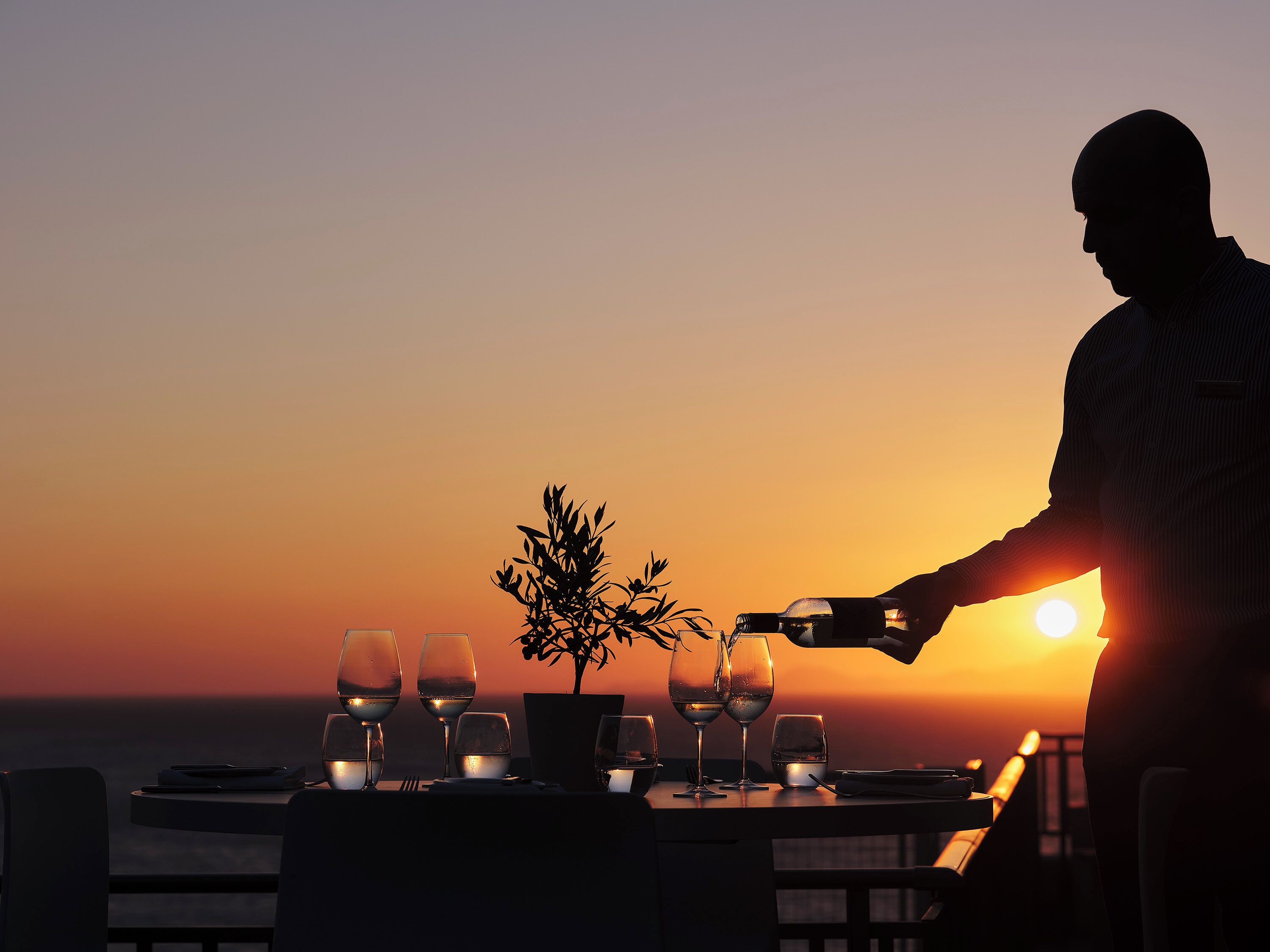Mesa preparada para una comida, con copas de vino y una planta, con vista a la puesta de sol.