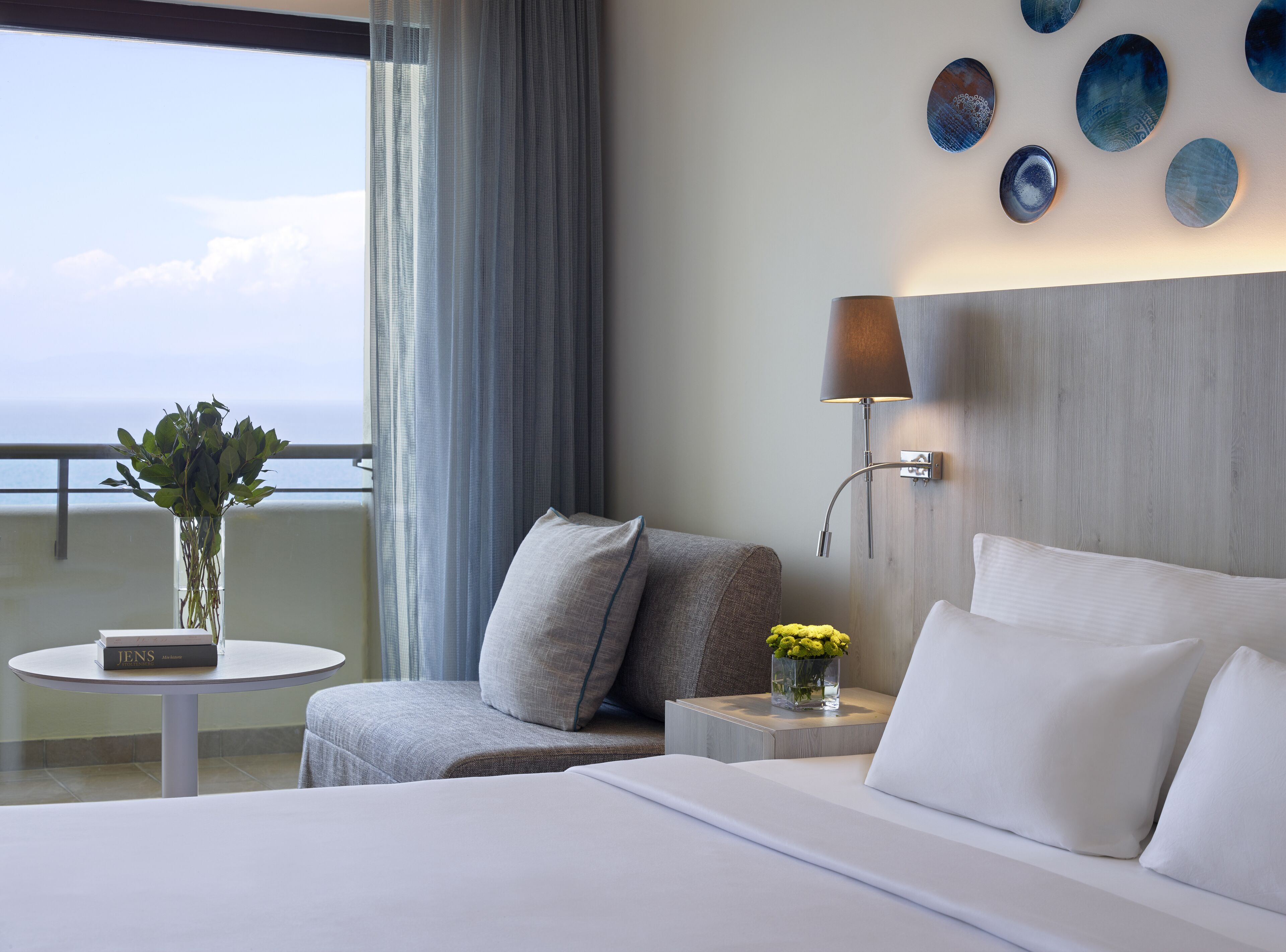 A hotel room with a bed, a nightstand, a lamp, a vase with flowers, a small table with a book, and a view of the ocean through a window.