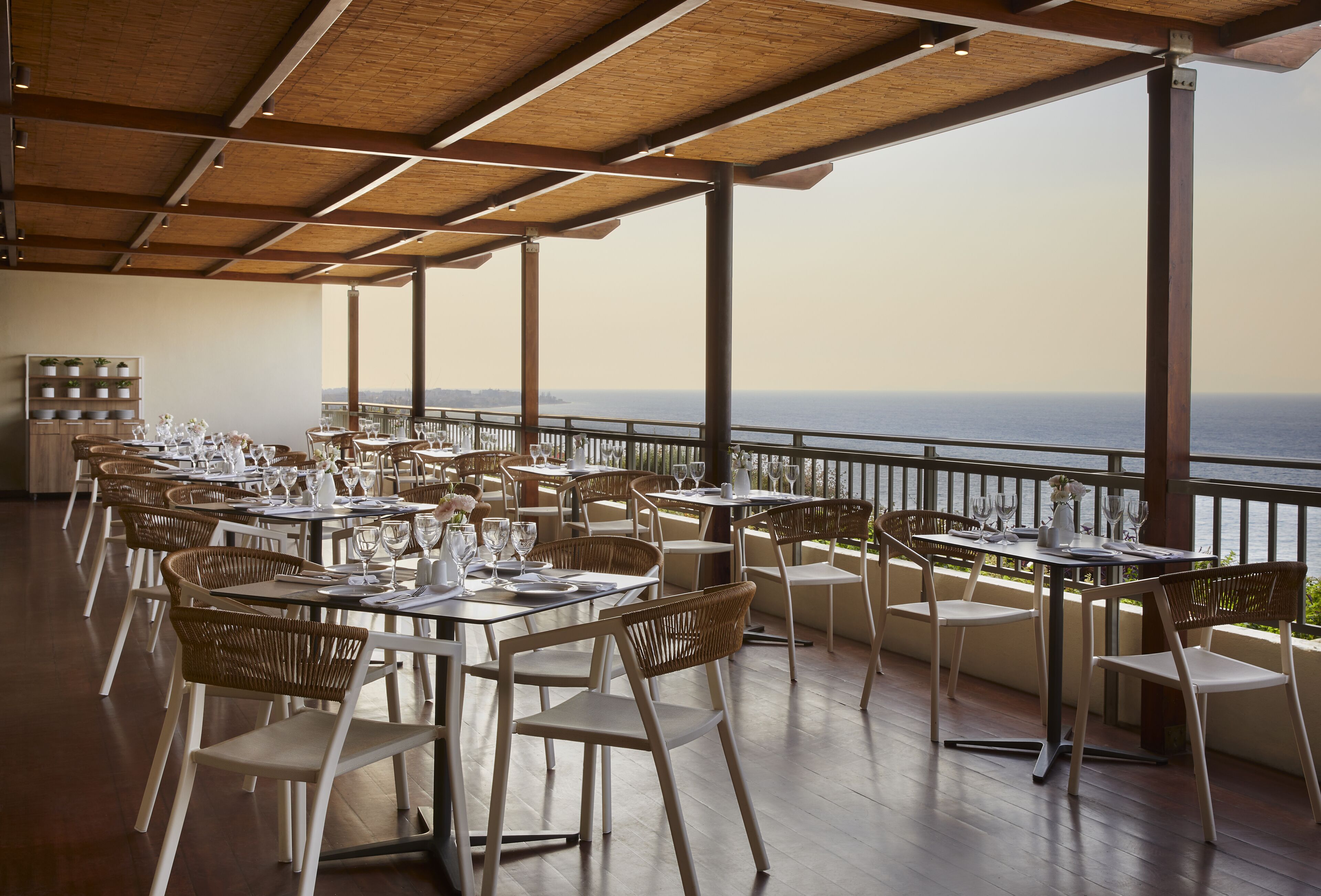 A restaurant with outdoor seating overlooking the sea, featuring wooden tables and wicker chairs.