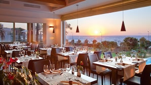 A restaurant with tables set for dining, large windows offering a view of the sea, and pendant lighting. - Amus Hotel & Spa (Rhodes)