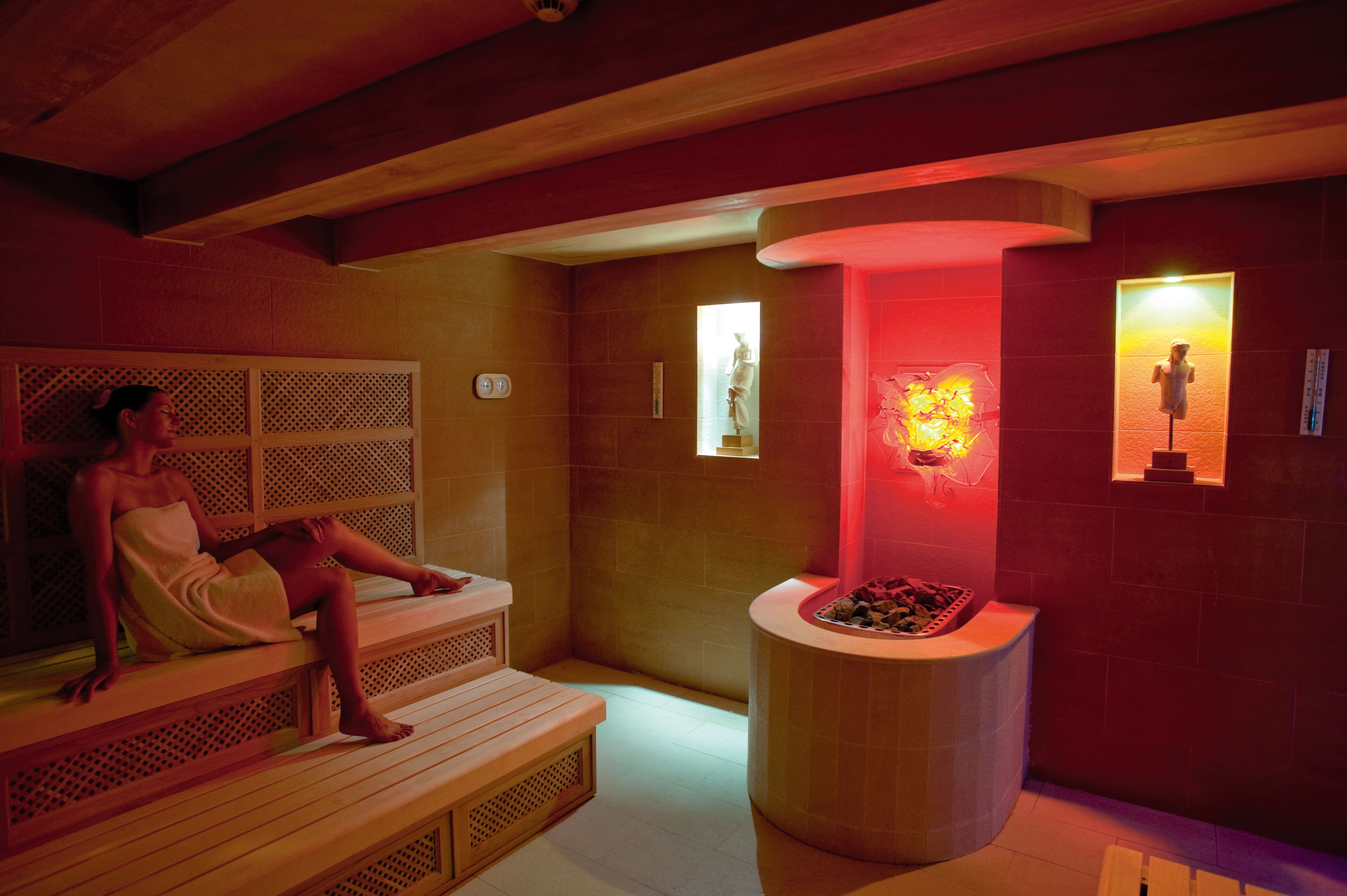 A sauna room with wooden benches, a central stone basin, and wall-mounted statues.