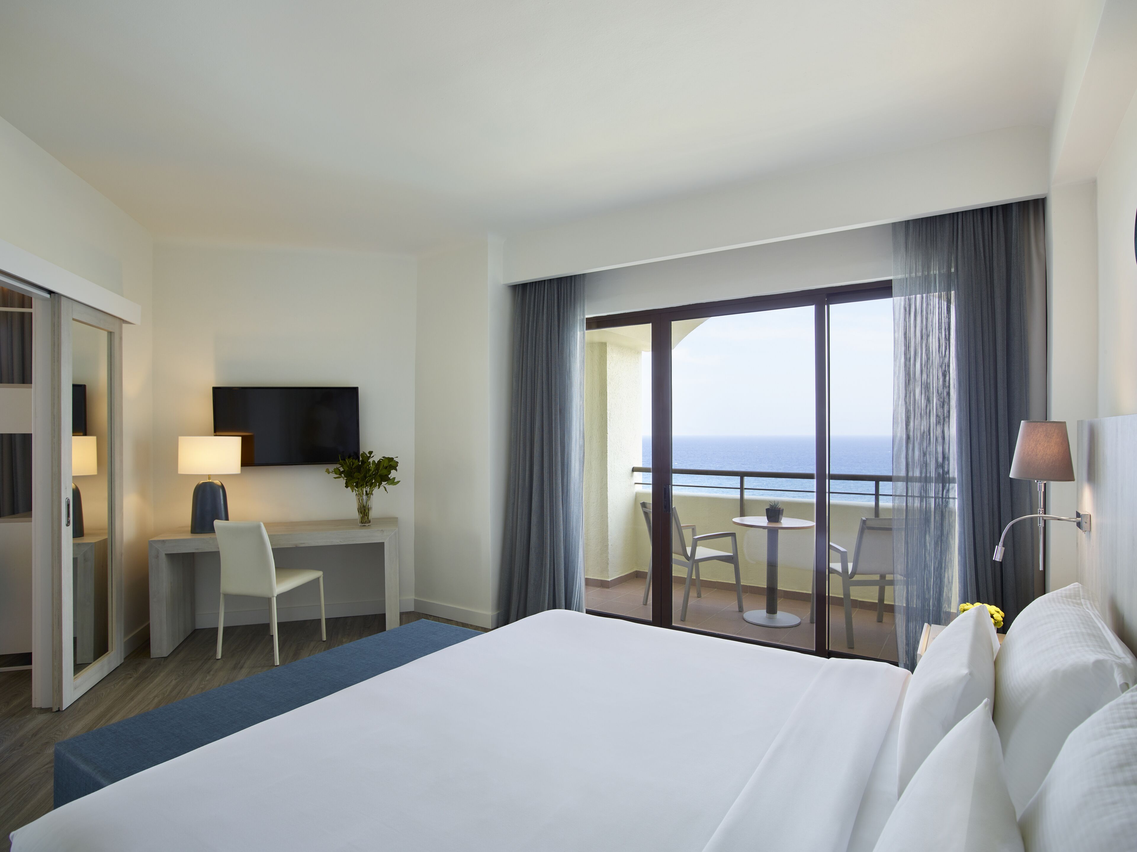 Habitación de hotel con cama, escritorio, silla, televisor y balcón con vista al océano.