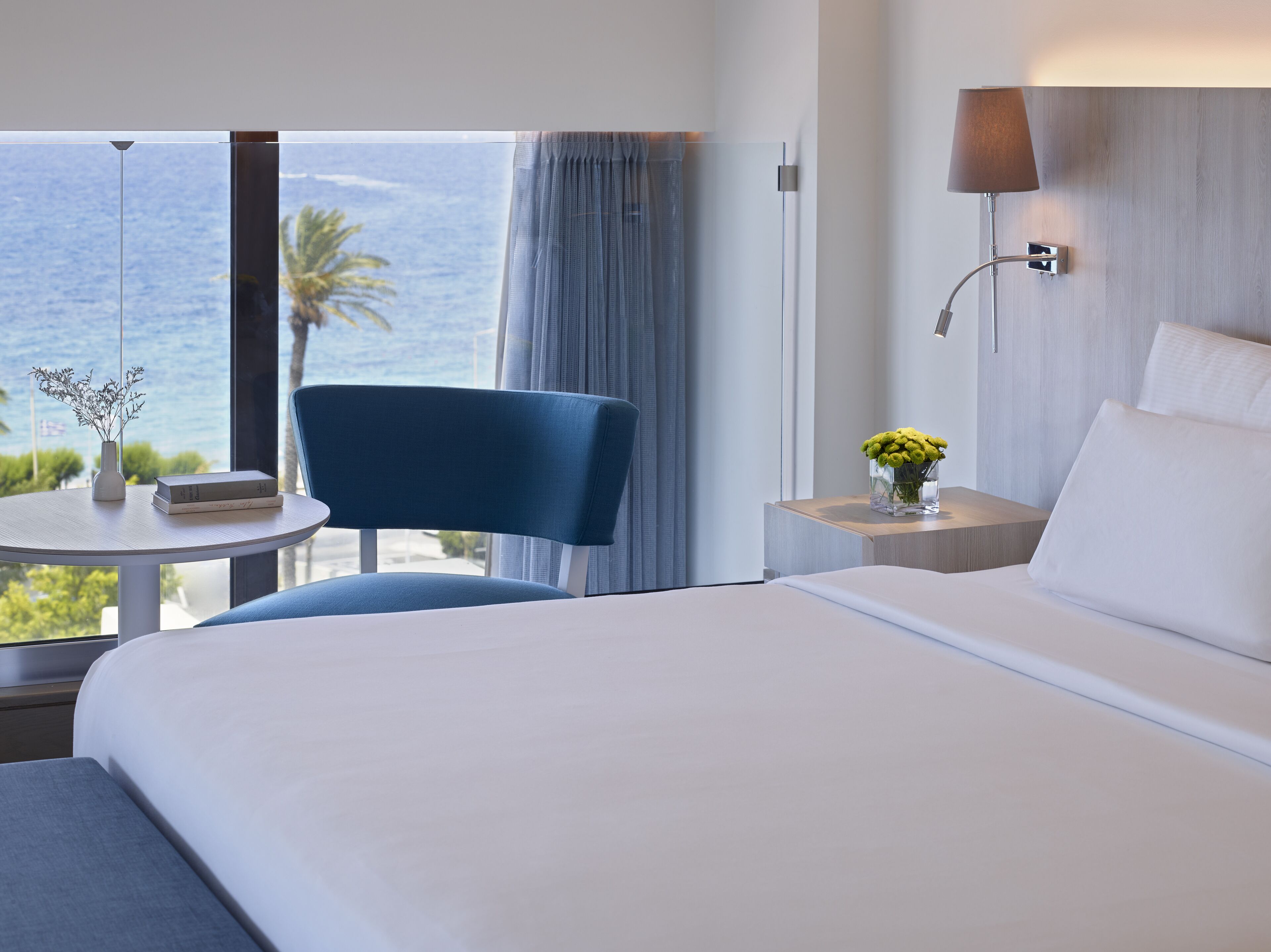 Habitación de hotel con cama, una silla, una mesita con un florero y vista al océano.