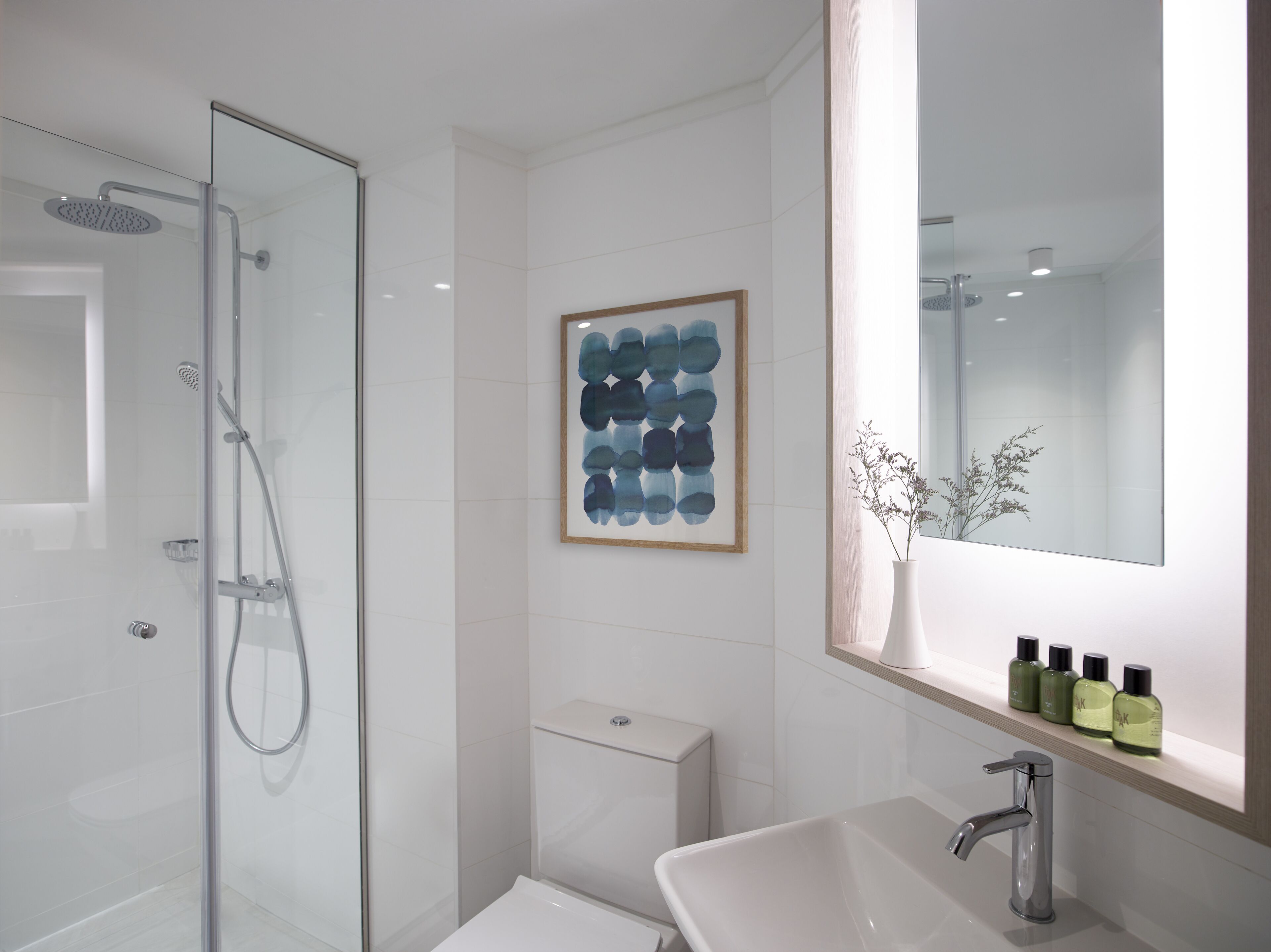 Un baño moderno con ducha envuelta en vidrio, inodoro, lavabo con espejo y un cuadro abstracto en la pared.