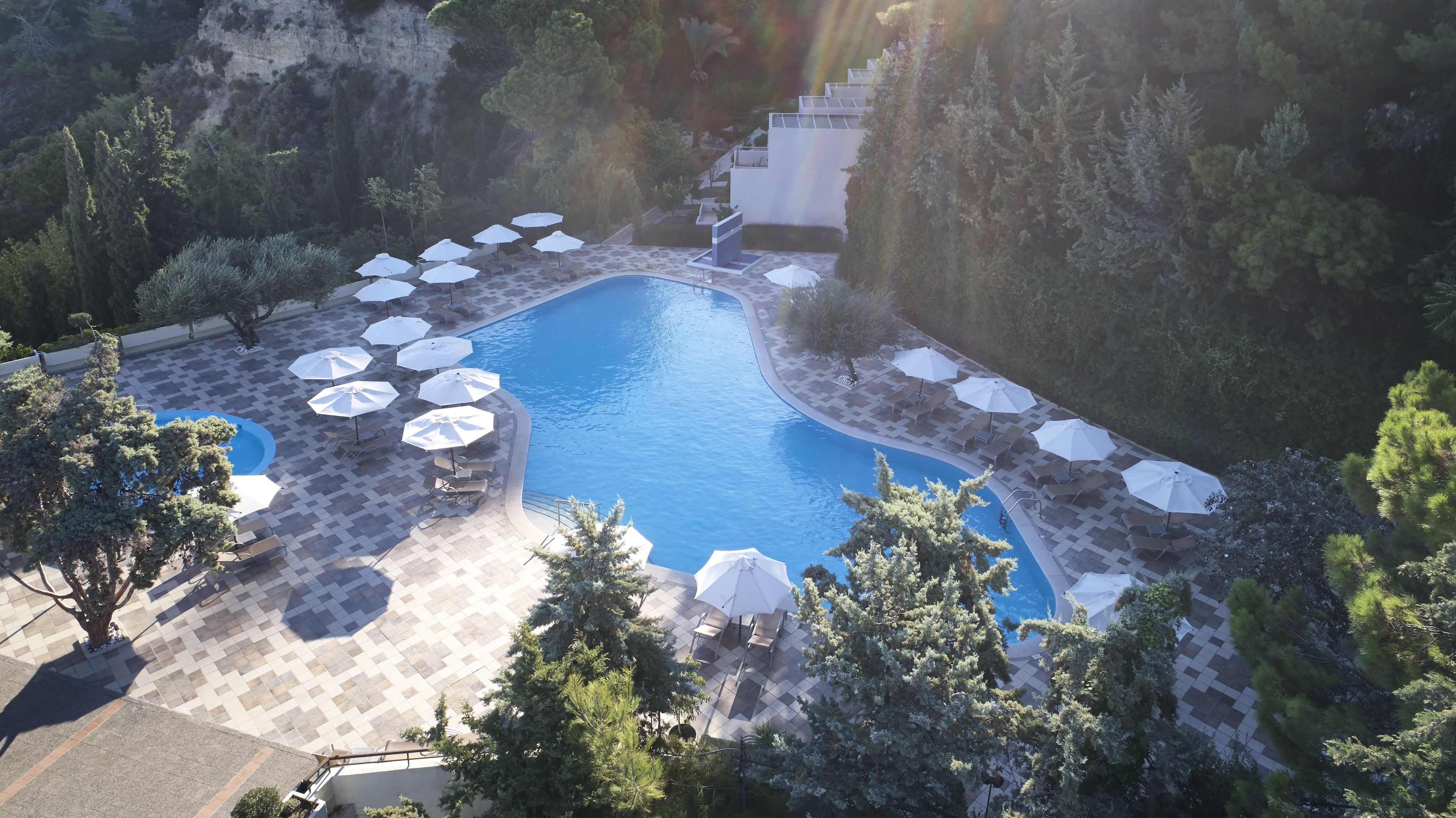 A pool area with multiple sun loungers and umbrellas, surrounded by trees and a mountainous backdrop.