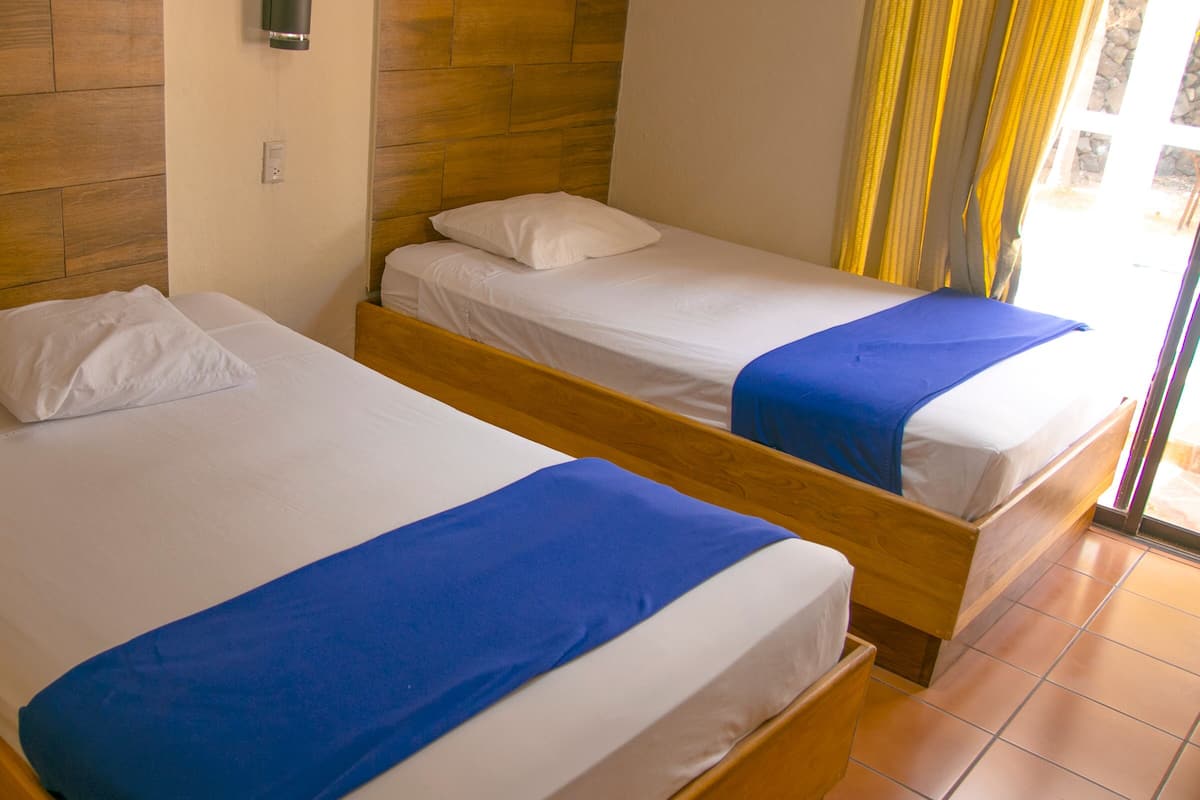 Duas camas de solteiro com cabeceiras de madeira, roupa de cama branca e colchas azuis em um quarto com paredes de madeira e piso ladrilhado.