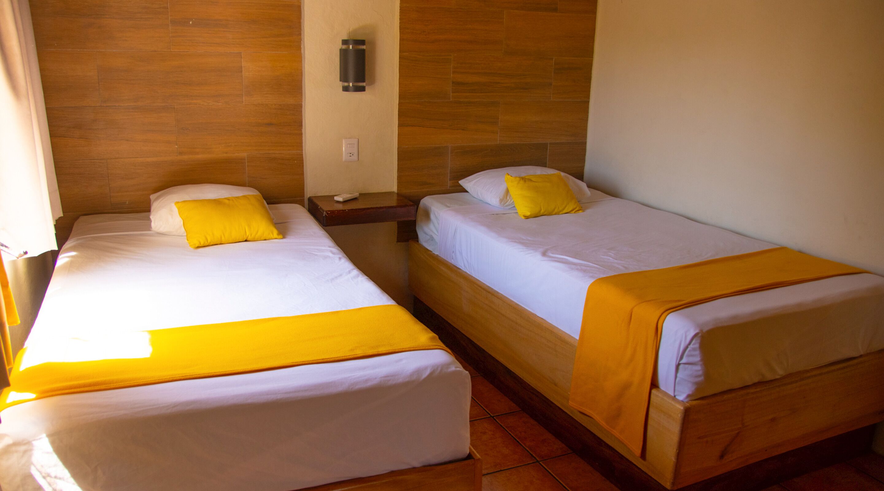 Two single beds with yellow pillows and blankets, wooden headboards, a nightstand with a lamp, and a wall-mounted heater.