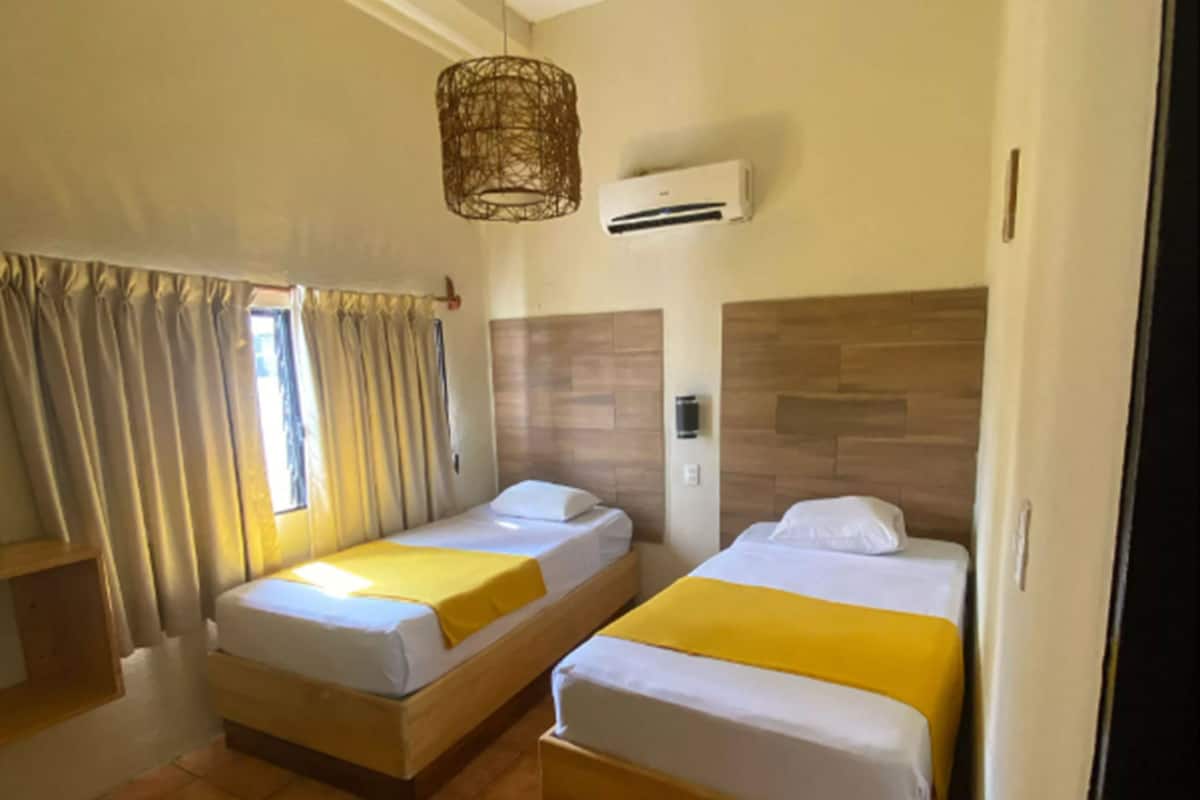 Quarto de hotel com duas camas, janela com cortinas, ar condicionado e luminária suspensa.