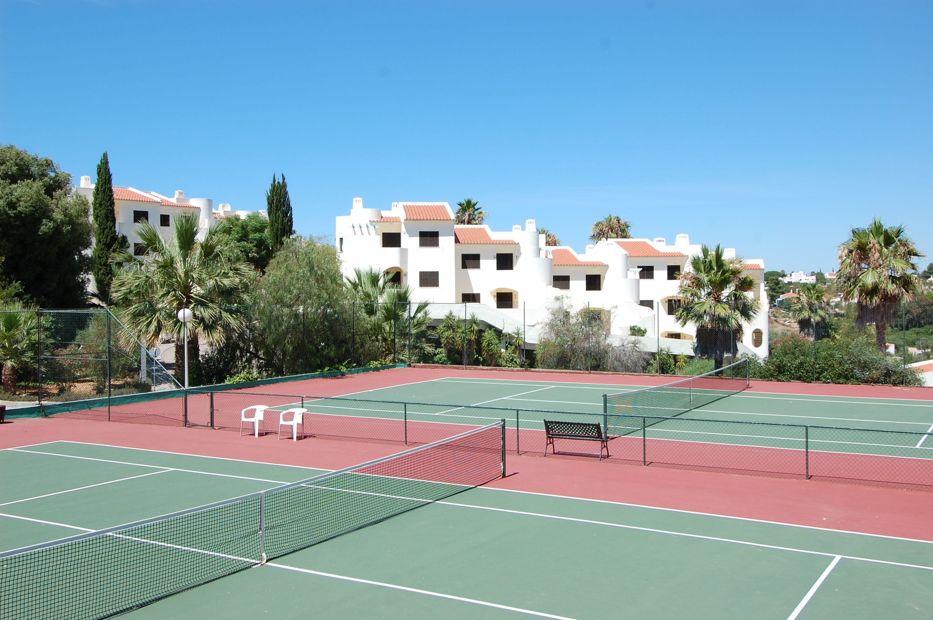 Tenniskenttä, jossa on verkko, kaksi valkoista tuolia ja penkki.