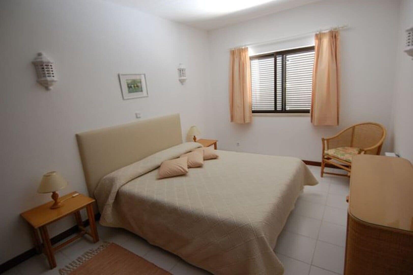 A bedroom with a bed, bedside tables, a chair, a window with curtains, and a wall-mounted light fixture.