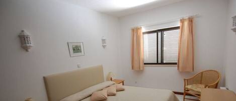 A bedroom with a bed, bedside tables, a chair, a window with curtains, and a wall-mounted light fixture.