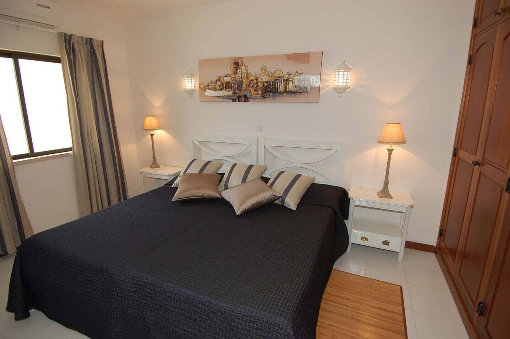 A neatly arranged bedroom with a large bed, two bedside tables with lamps, a wall-mounted artwork, and a wooden wardrobe.