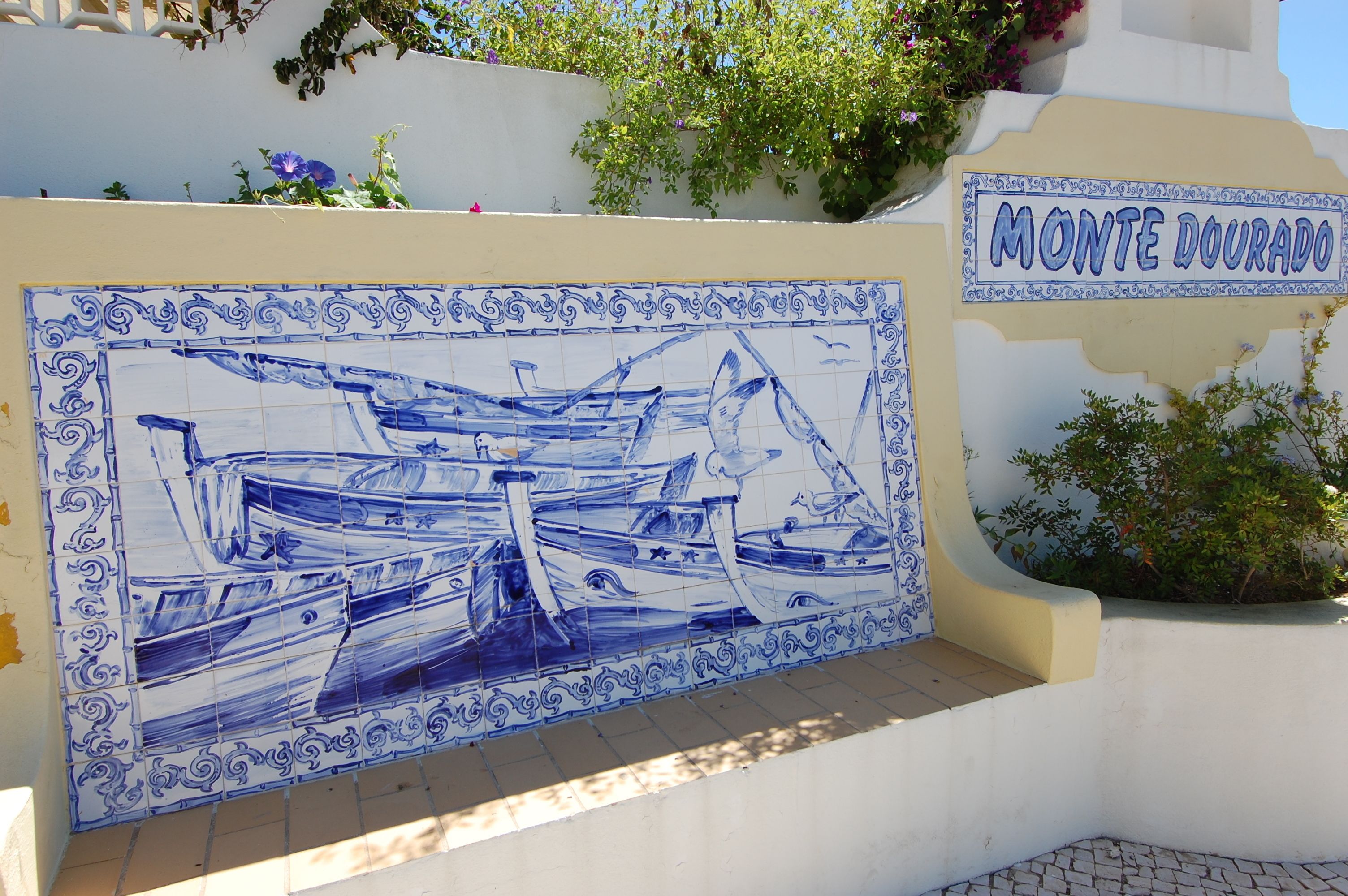 Eine mit blauen und weißen Fliesen verkleidete Wand, die eine maritime Szene zeigt, mit einem Schild mit der Aufschrift 'Monte Dourado'.