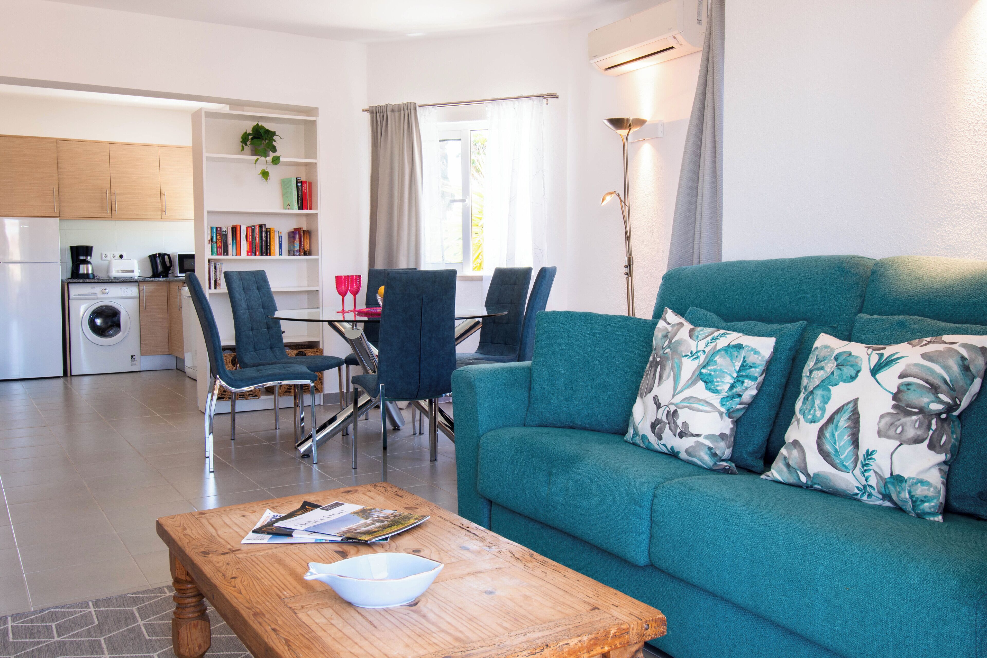 Una sala moderna con un sofá verde azulado, una mesa de centro de madera, y un comedor con sillas y una mesa.