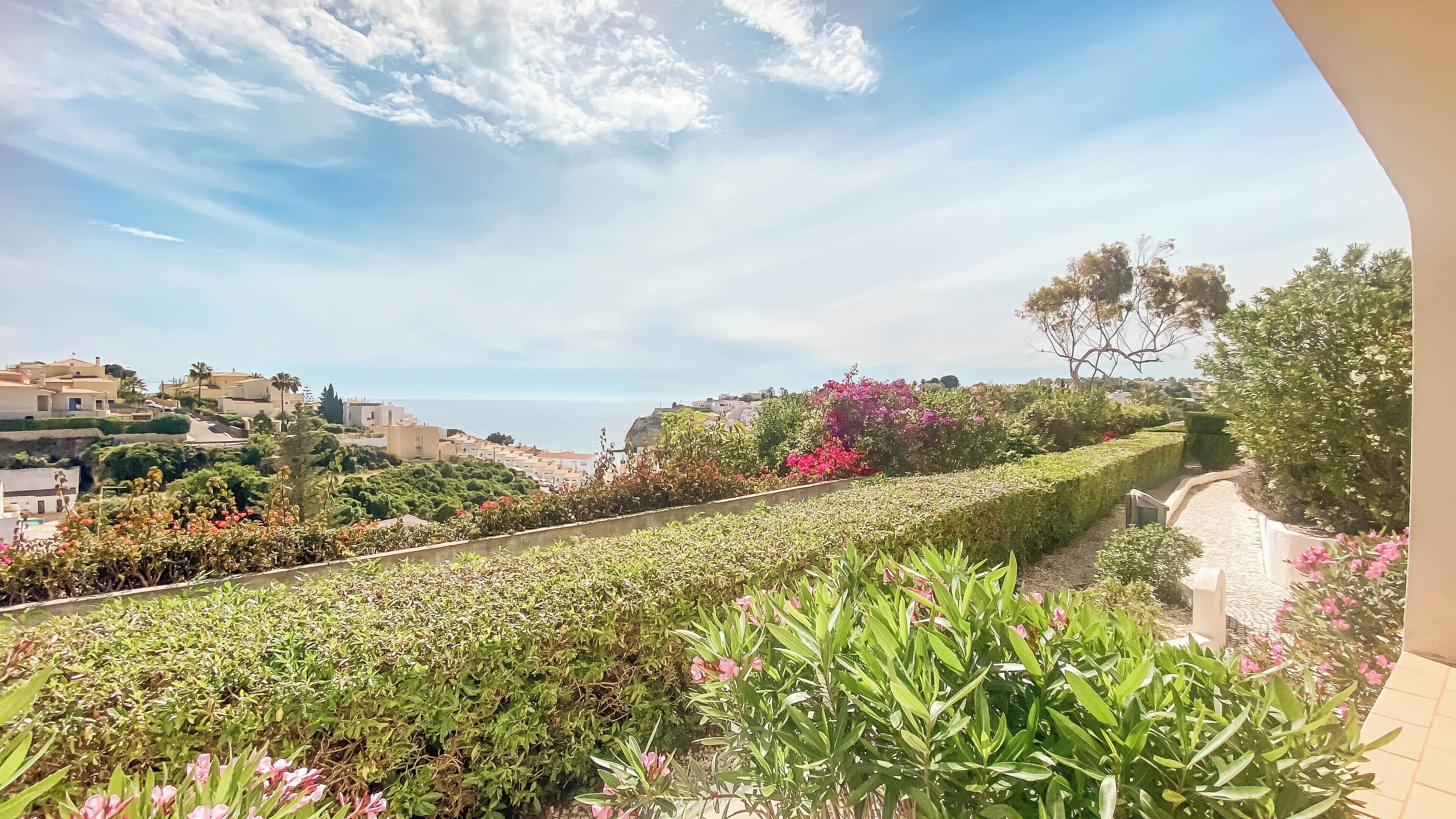 Un jardín ajardinado con diversas plantas y flores, un sendero y una vista de un pueblo costero con edificios y el mar al fondo.