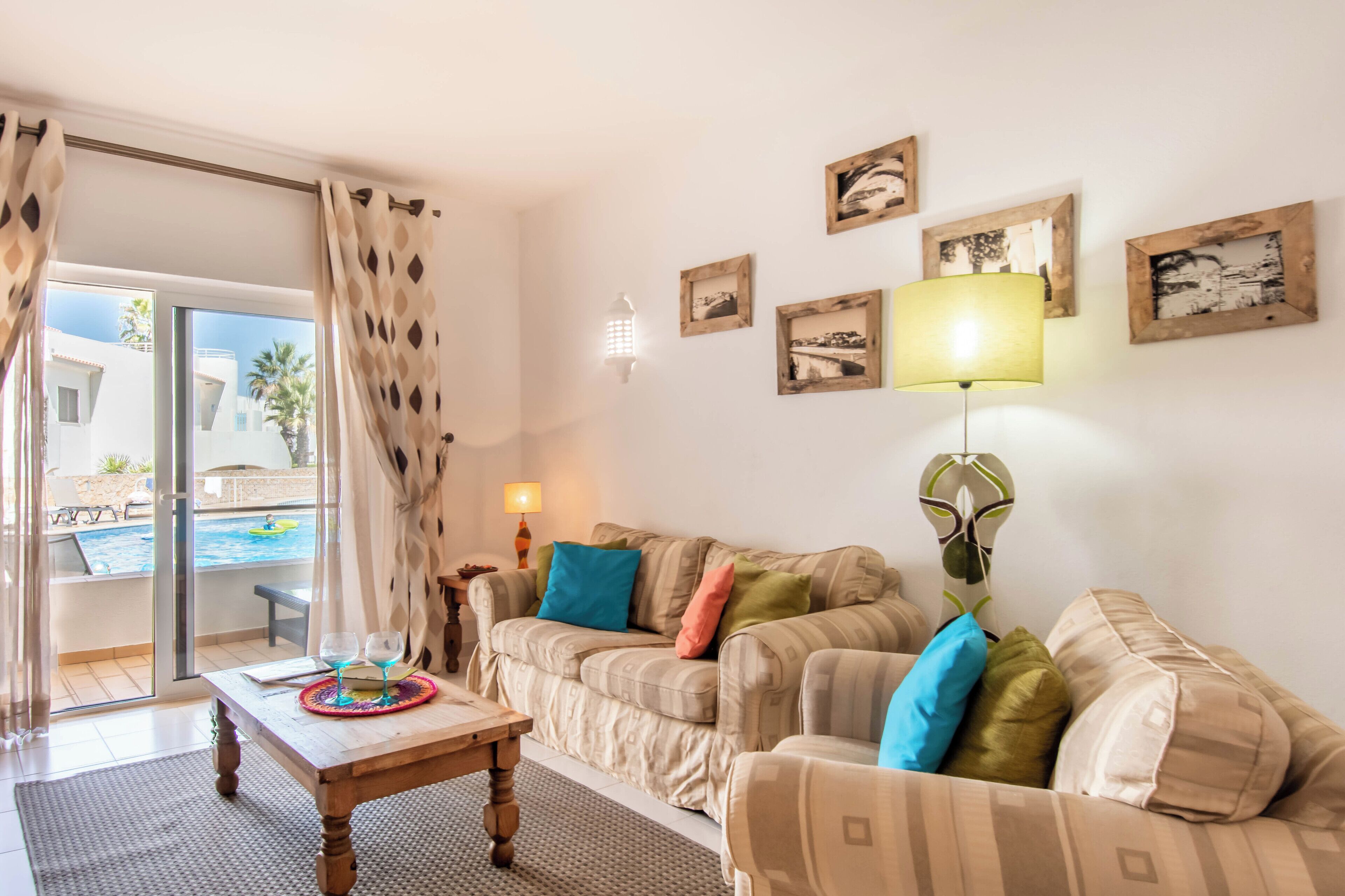 Un salon comprenant un canapé, une table basse et une lampe. Les murs sont ornés de tableaux encadrés, et une fenêtre donne sur une piscine.