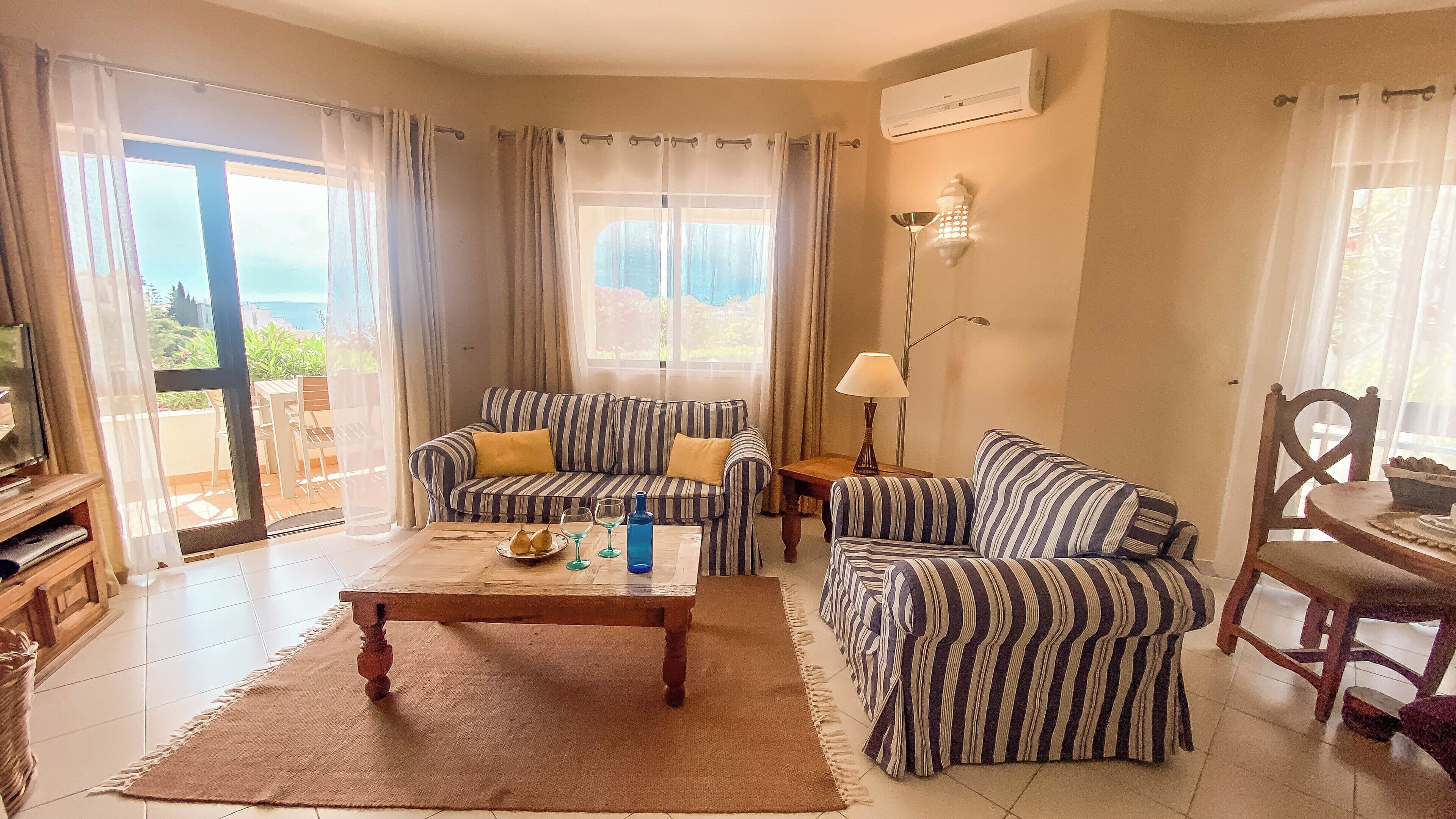 A living room with a sofa, armchair, coffee table, and a TV. There is a window with curtains, a lamp, and an air conditioning unit on the wall.