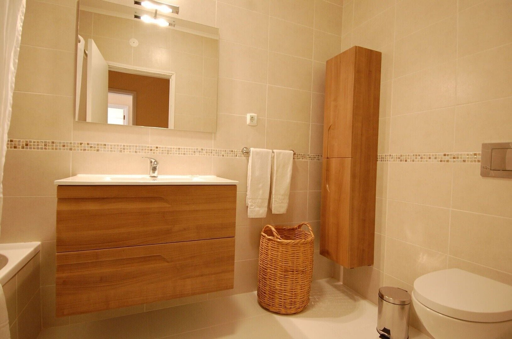 Un baño con tocador de madera, espejo, lavamanos, inodoro y cesta de mimbre.