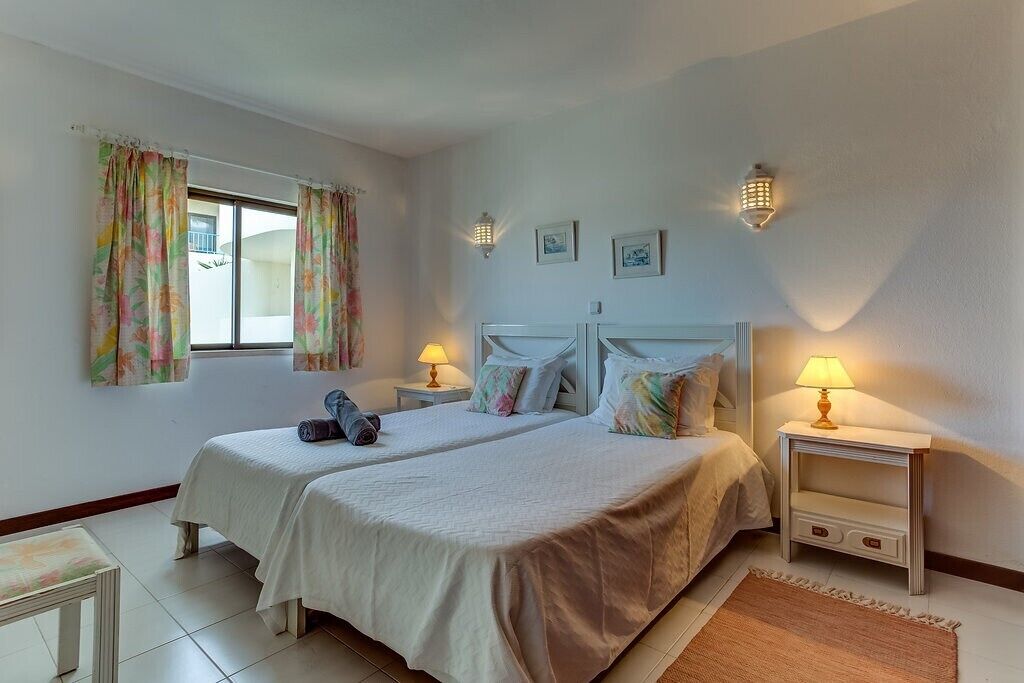 Un lit double recouvert d’une courtepointe blanche, deux tables de chevet avec des lampes, une fenêtre ornée de rideaux à motifs floraux et une petite table avec une chaise.