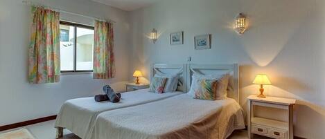 A double bed with a white bedspread, two bedside tables with lamps, a window with floral curtains, and a small table with a chair.