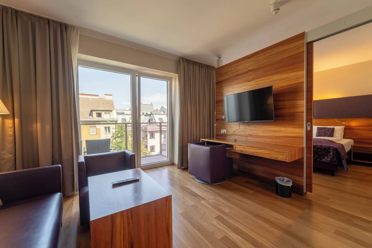 A modern hotel room with a wooden accent wall, a flat-screen TV, a sofa, a dining table, and a view of the city through large windows.