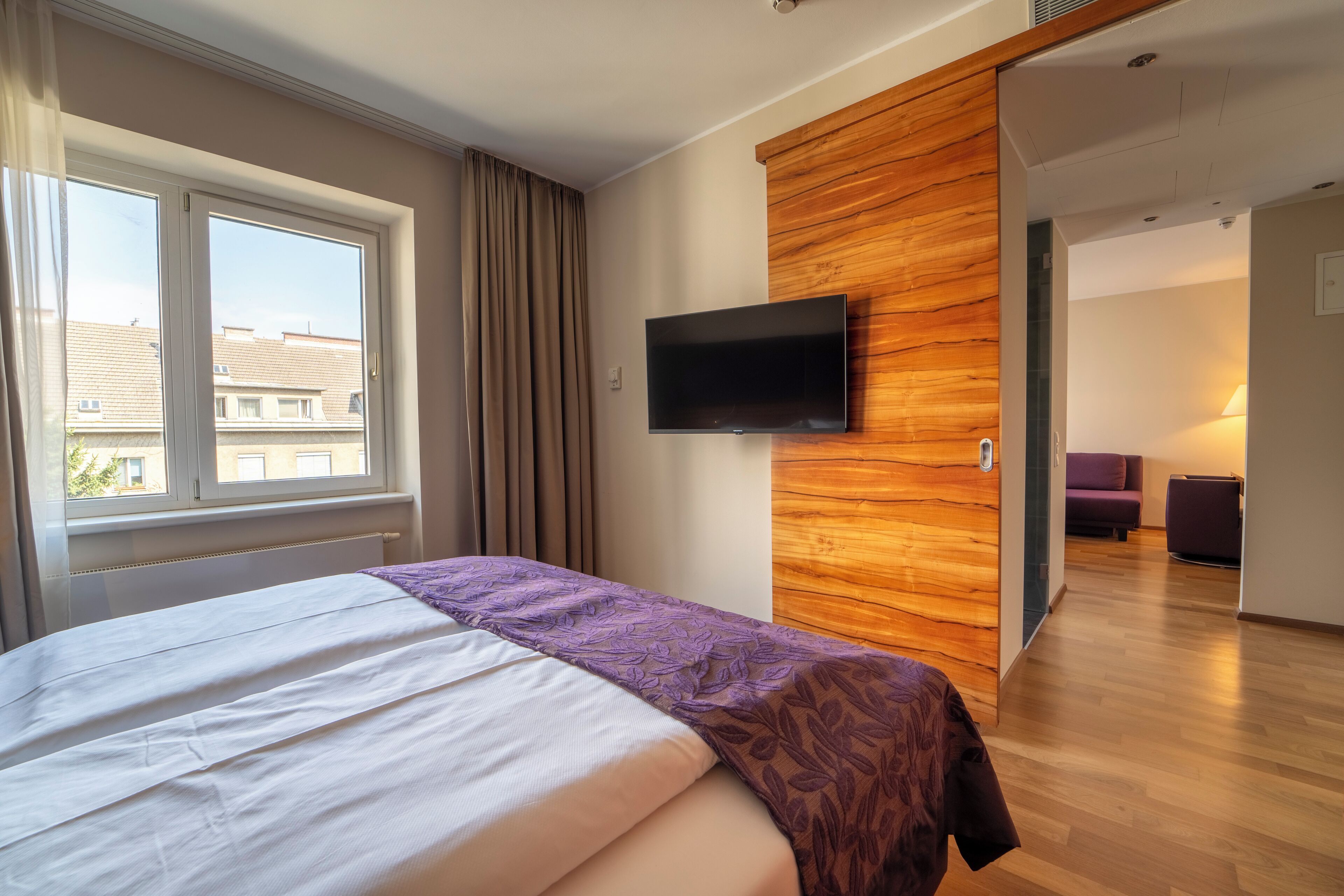 A modern hotel room with a large bed, a TV mounted on a wooden wall, and a view of the city through the window.
