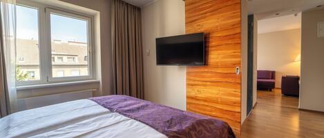 A modern hotel room with a large bed, a TV mounted on a wooden wall, and a view of the city through the window.