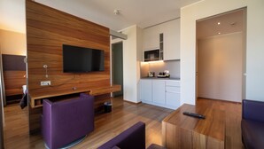 A modern living room with a wooden accent wall, a flat-screen TV, purple seating, and a kitchen area with white cabinets and a microwave. - Pakat Suites (Vienna)
