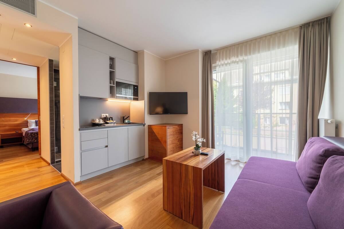 A modern hotel room with a wooden floor, a purple sofa, a wooden coffee table, a flat-screen TV mounted on the wall, and a kitchen area with white cabinets and a microwave.