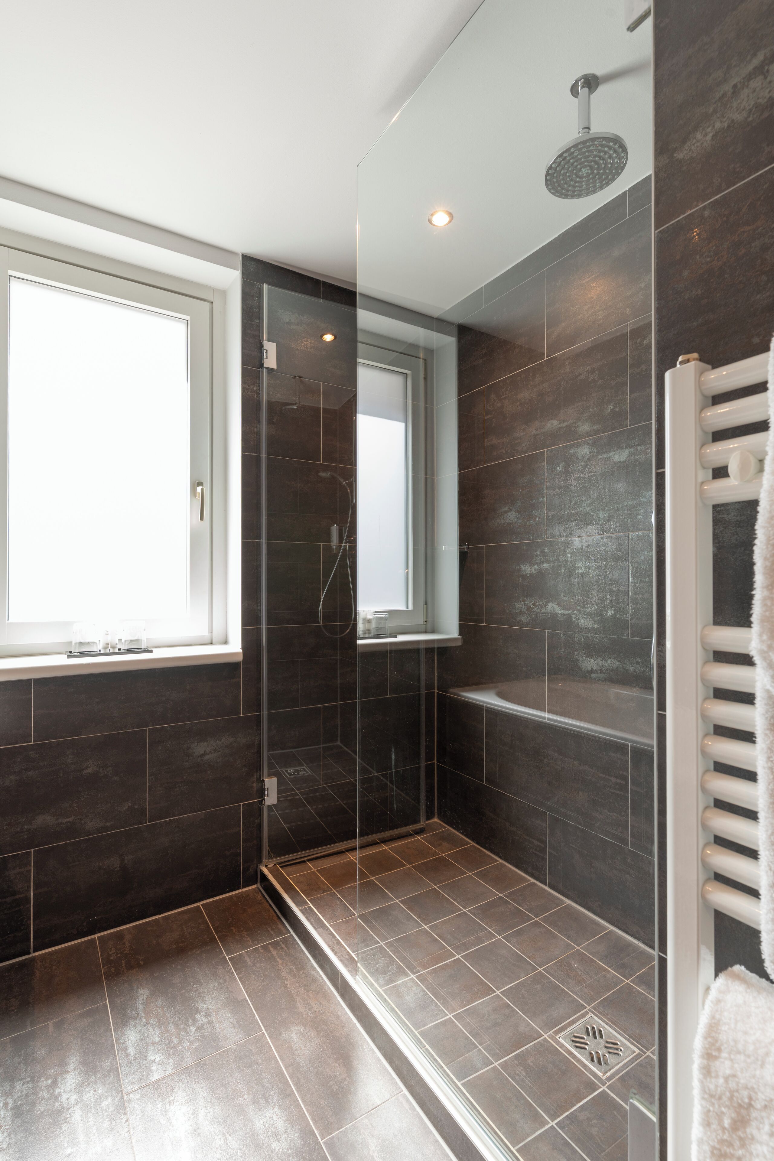 Een moderne badkamer met een douchecabine van glas, donkere tegels, een raam en een witte handdoekhouder.