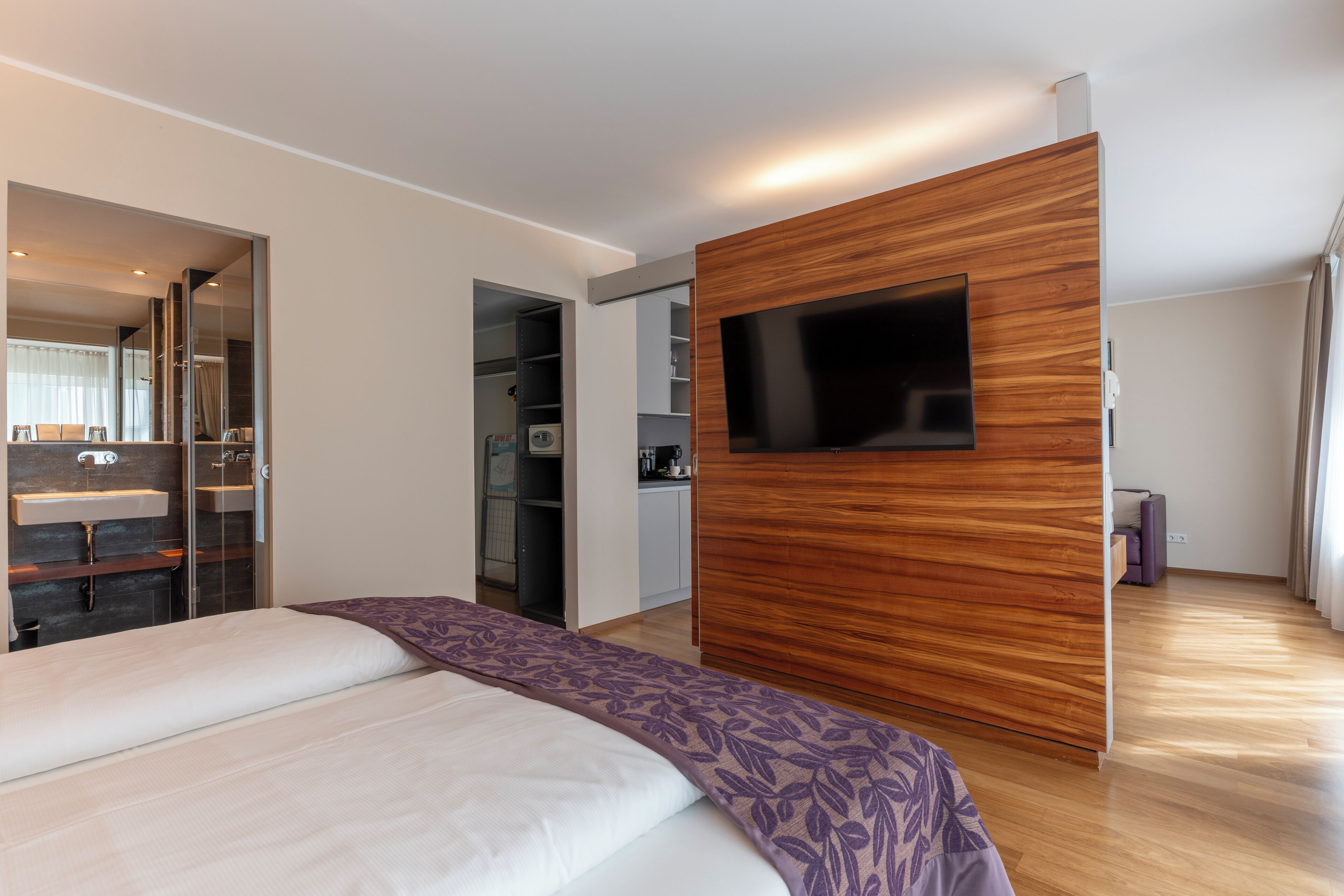A modern hotel room with a large bed, a wooden panel wall, a flat-screen TV, and a bathroom with a glass shower enclosure.