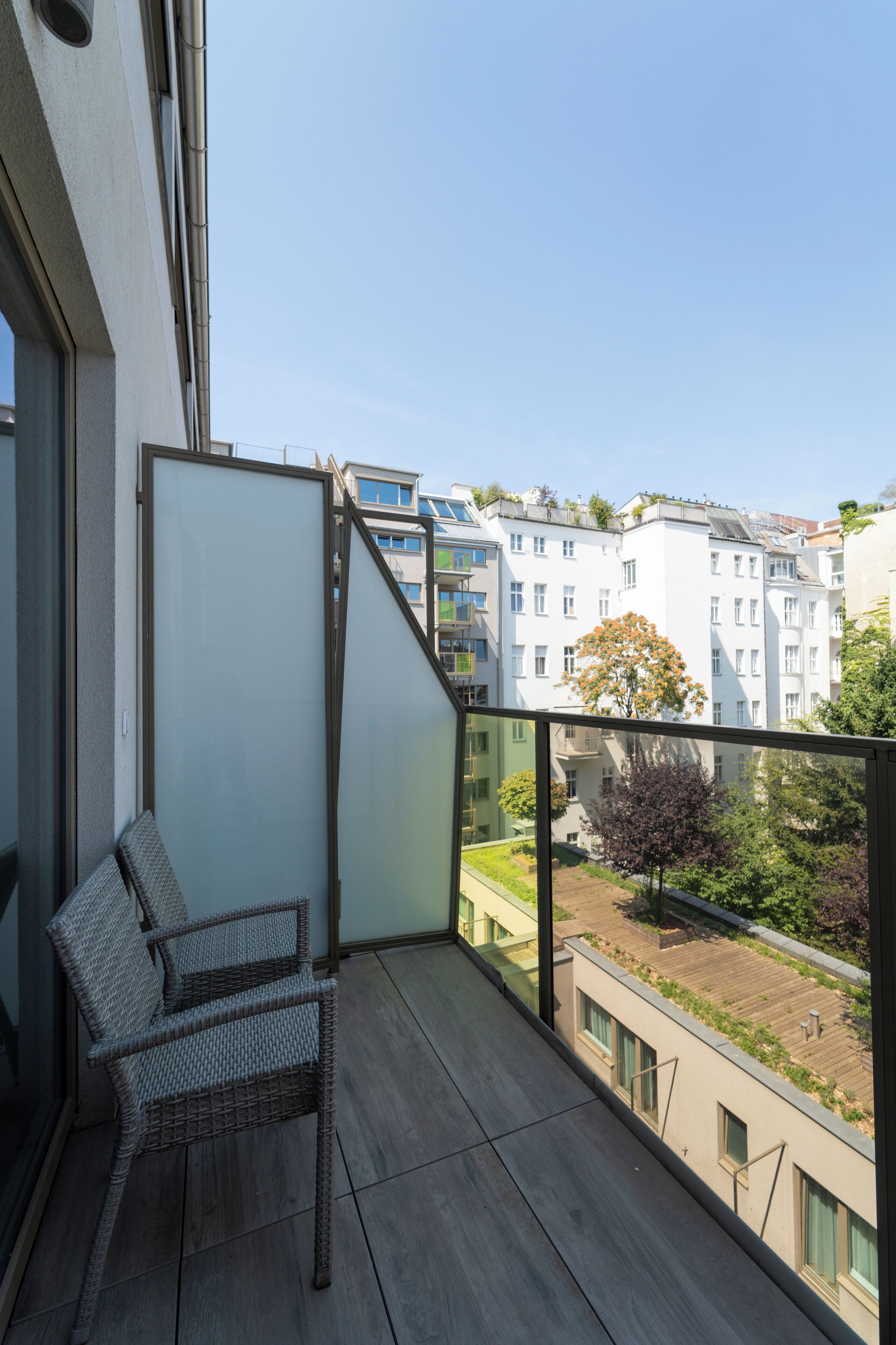 A balcony with a wicker chair, a frosted glass partition, and a view of residential buildings.