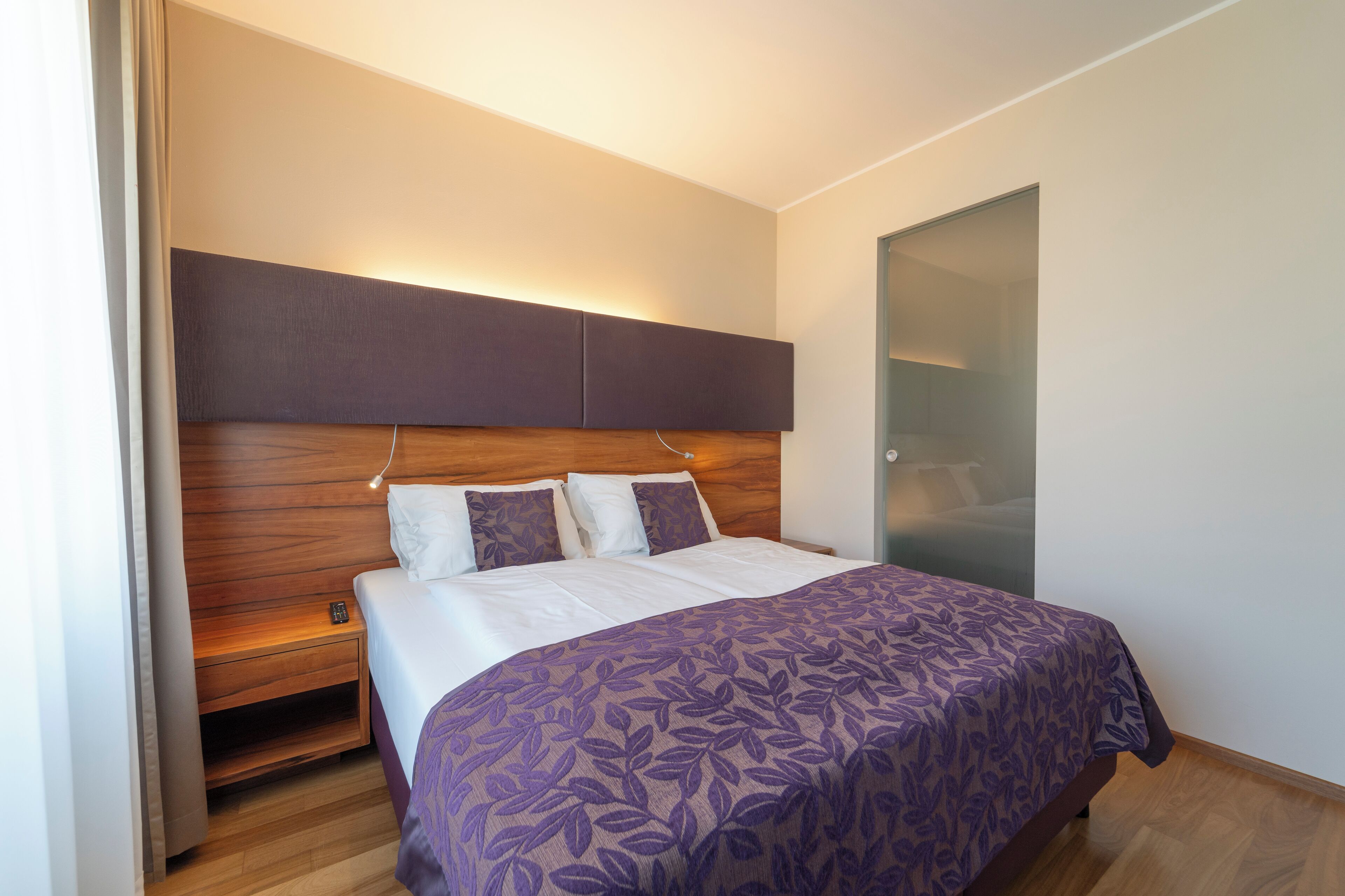 A modern hotel room with a large bed, wooden headboard, and a nightstand.