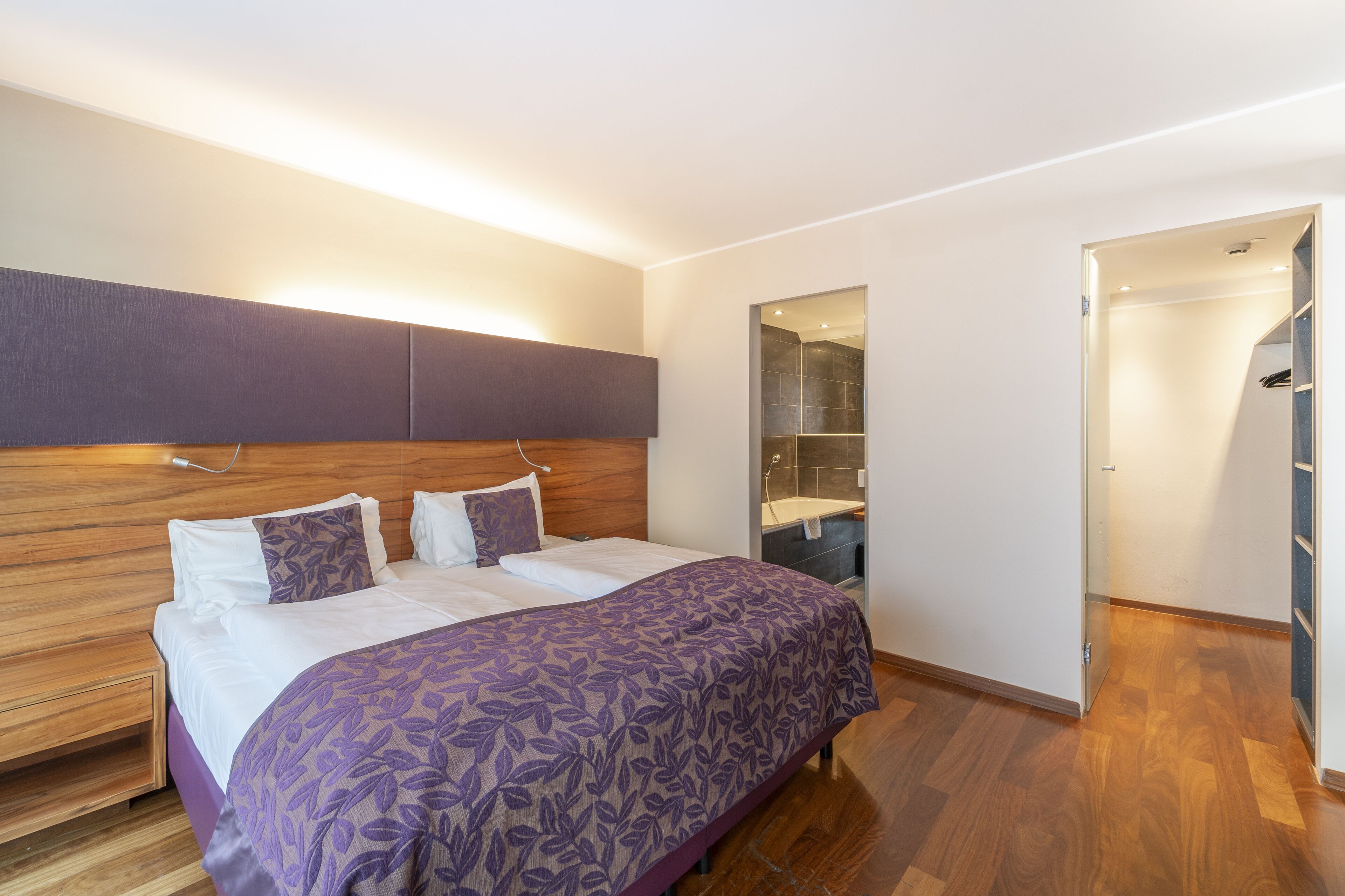 A modern bedroom with a wooden headboard, a large bed with a purple floral bedspread, a built-in bench, and a walk-in closet.