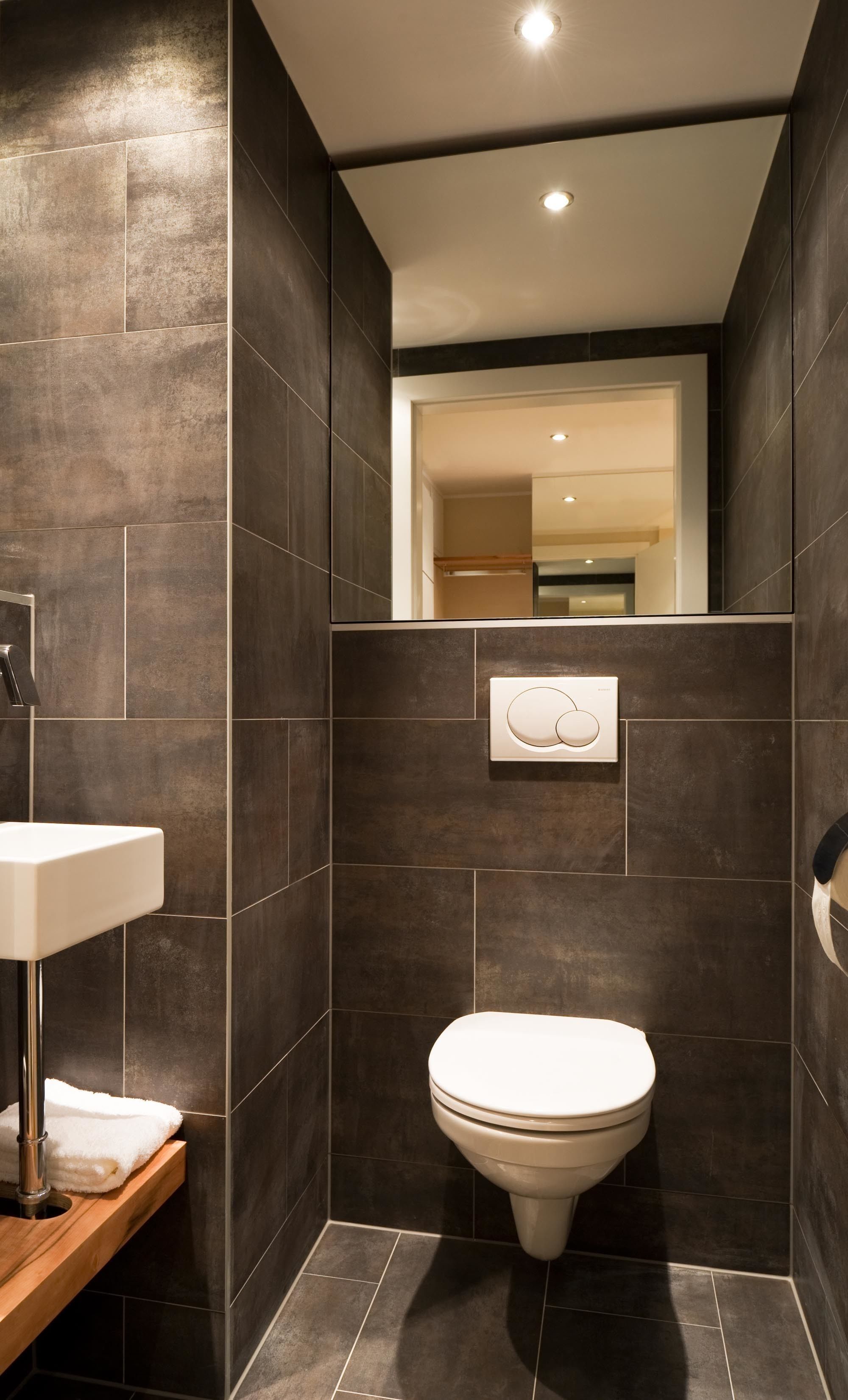 A modern bathroom with dark tiles, a white toilet, and a built-in sink with a towel.