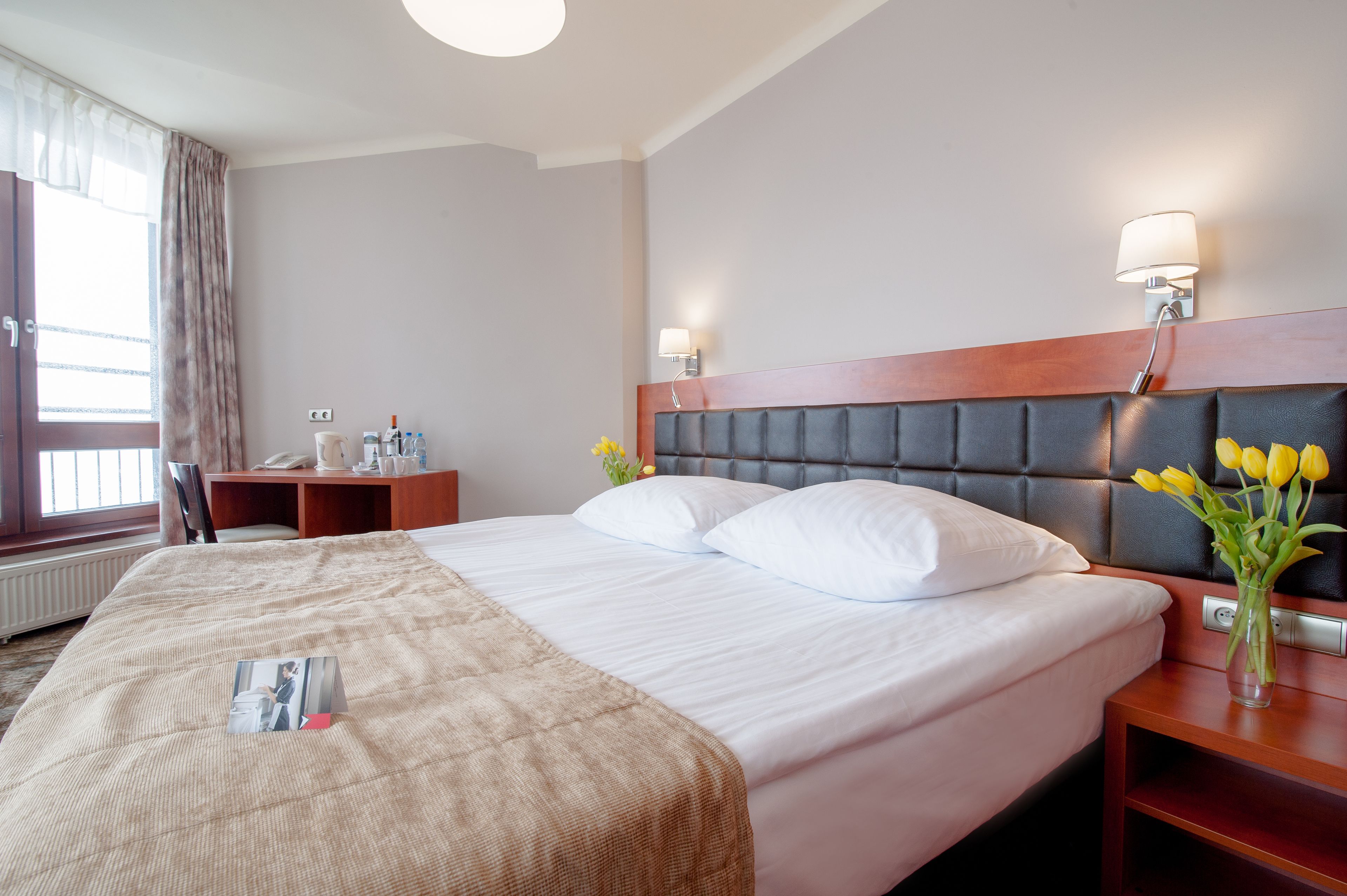 Una habitación de hotel con una cama grande, una mesita de noche con lámpara, un escritorio con silla y una ventana con cortinas.