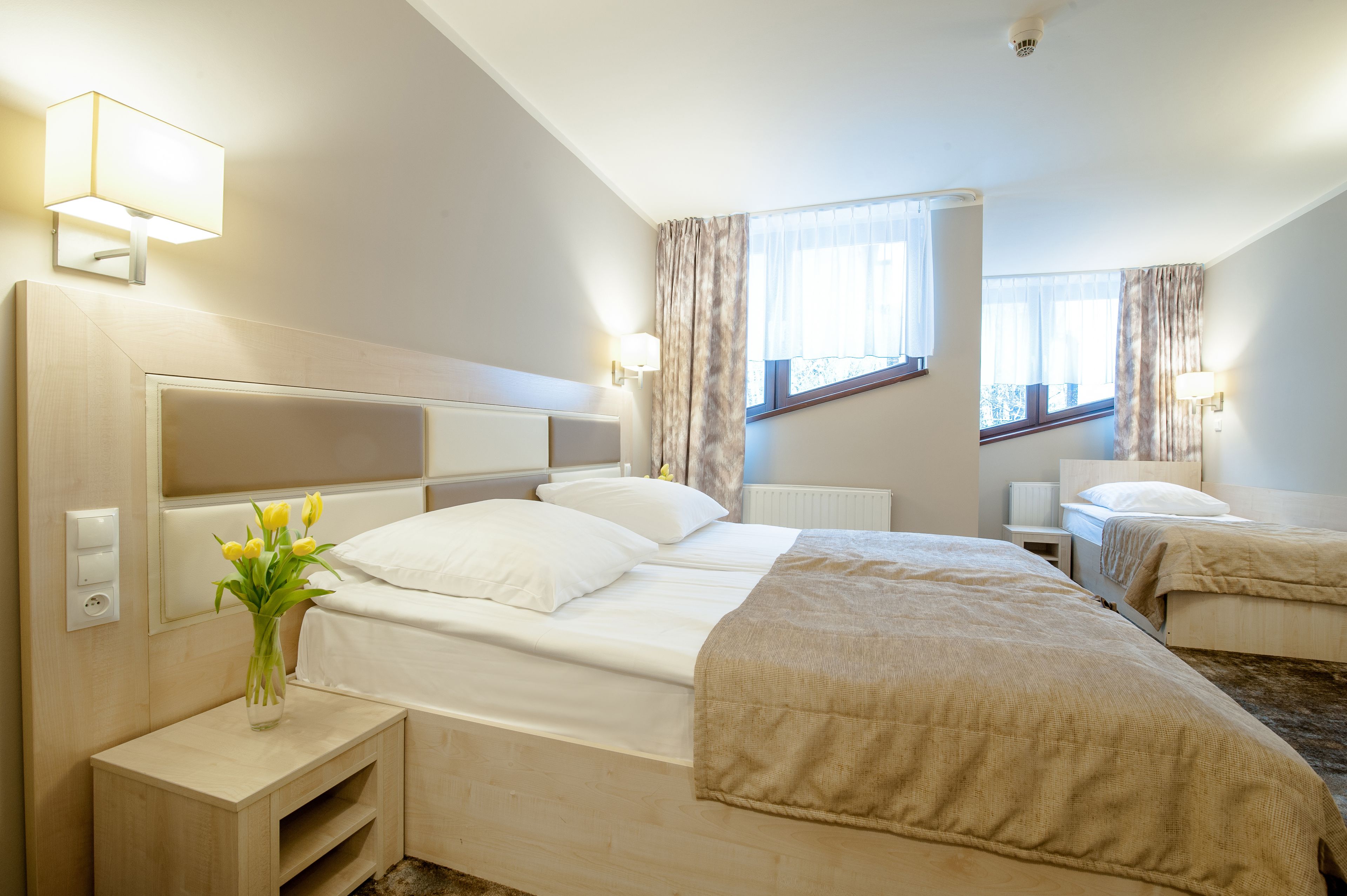 Una habitación de hotel con una cama grande, mesitas de noche, un jarrón con flores y dos ventanas con cortinas.