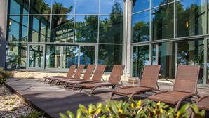 Une terrasse ensoleillée avec des fauteuils de jardin en osier, un mur de pierre et un bâtiment en verre en arrière-plan.