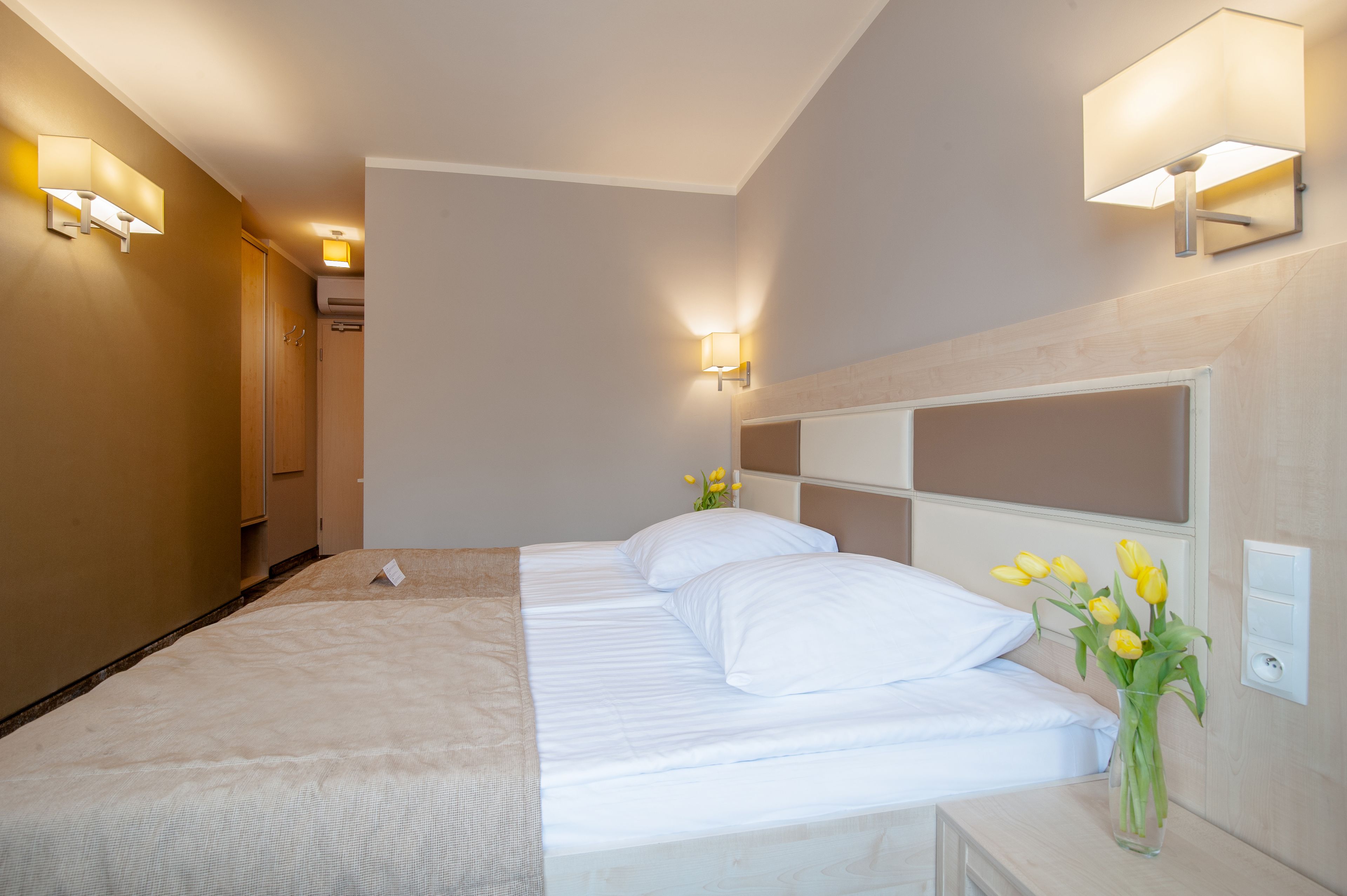 Una habitación de hotel con una cama, dos lámparas fijadas a la pared, un florero con flores y un interruptor de luz en la pared.