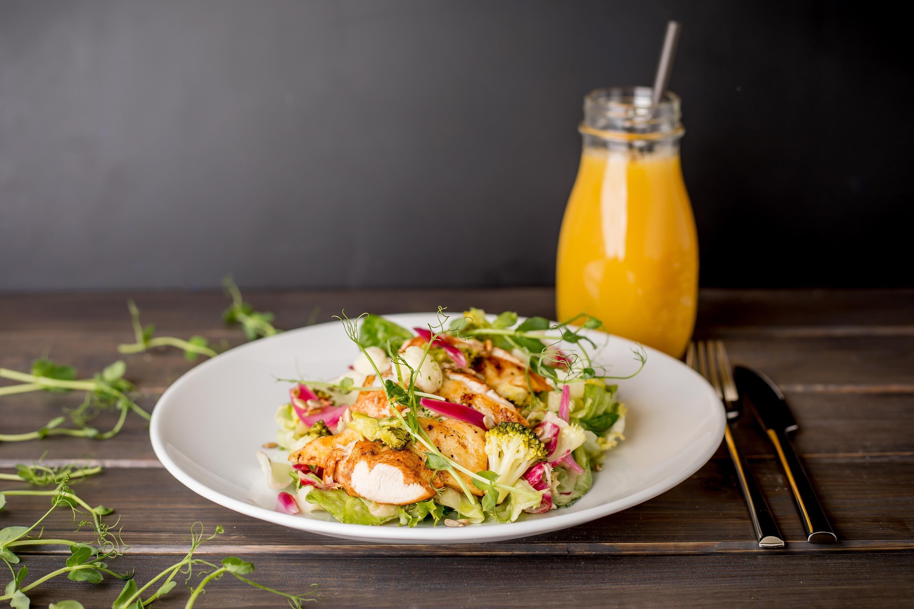 A plate of salad with chicken and vegetables, a glass of orange juice, and a fork and knife on a wooden table.