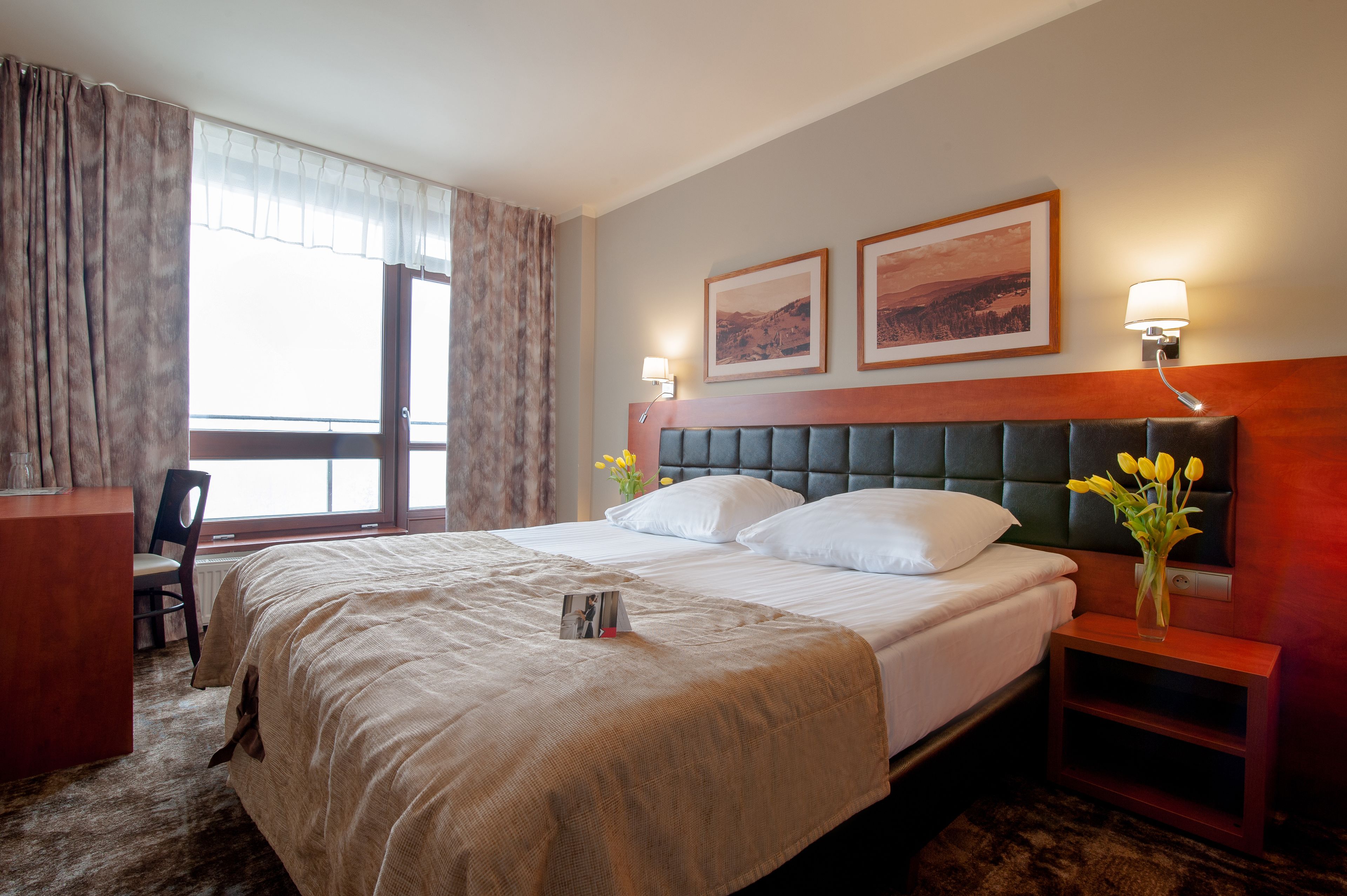 Une chambre d’hôtel avec un grand lit, un bureau et une chaise. Deux tableaux encadrés ornent le mur, et un vase rempli de fleurs est posé sur la table de chevet.