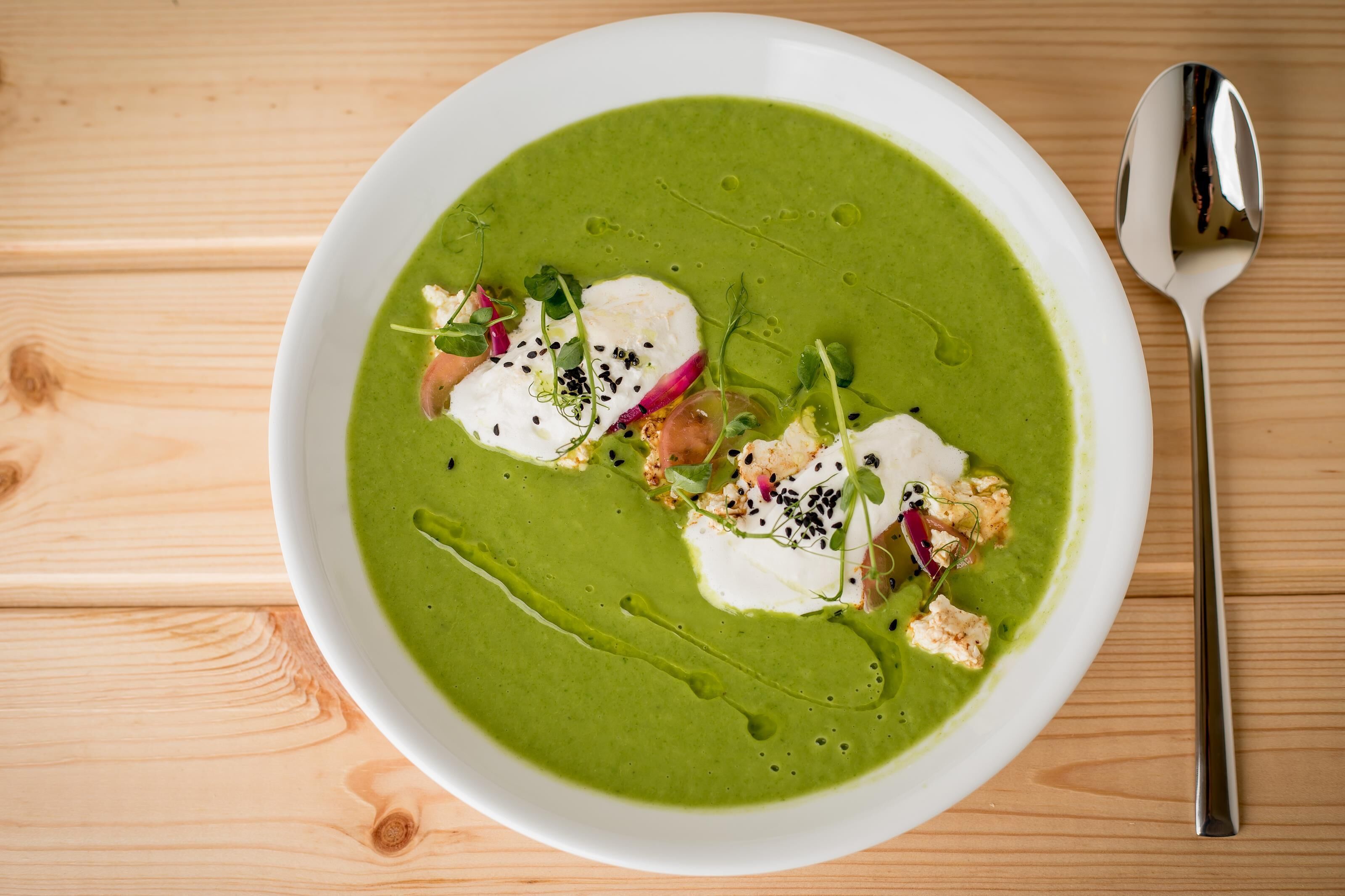 A bowl of green soup with white and red garnishes on a wooden table.